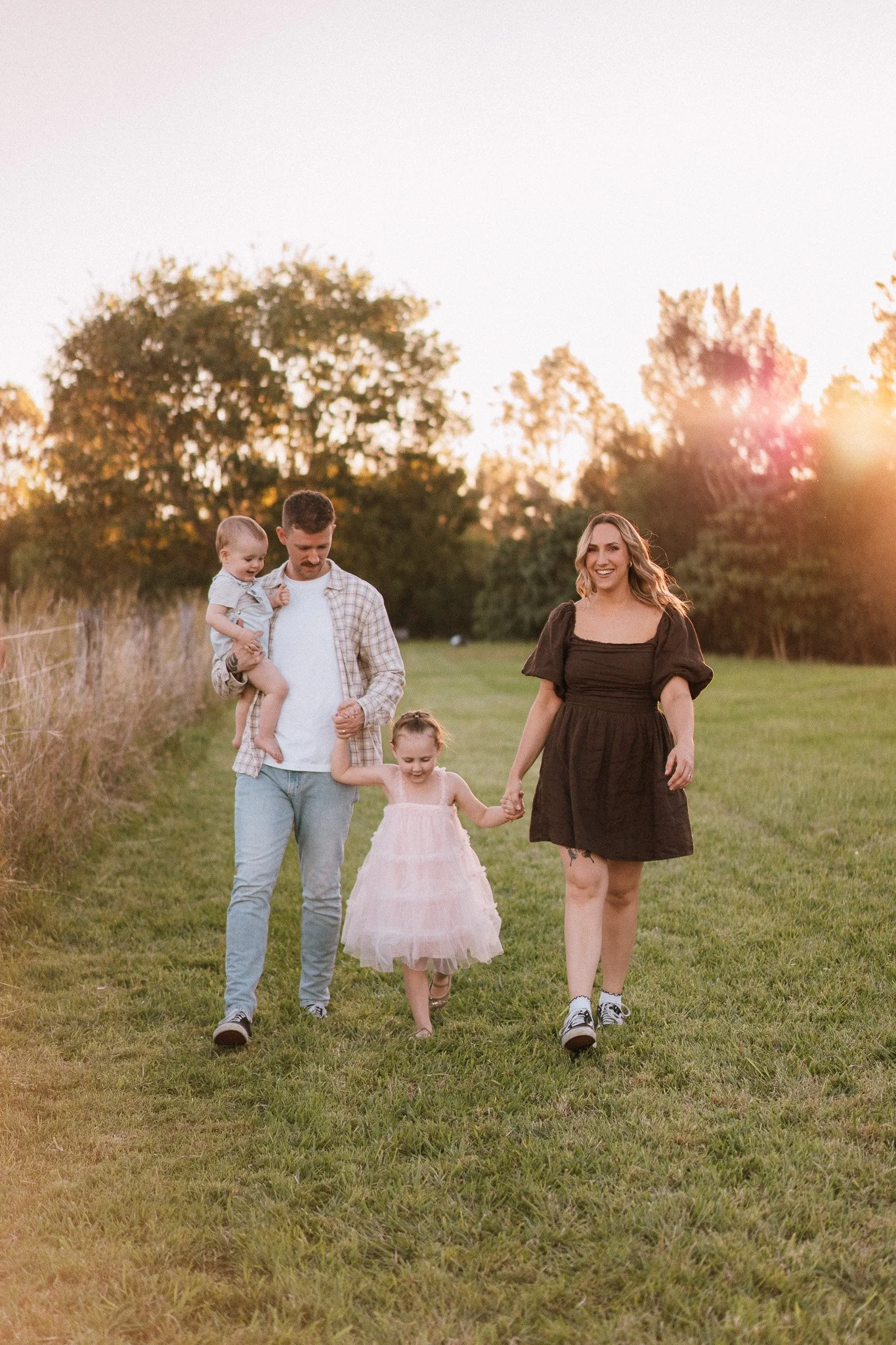 A family of four walking hand in hand on a grassy field during sunset. The father carries a young child, and a woman and a young girl are walking beside them, smiling.