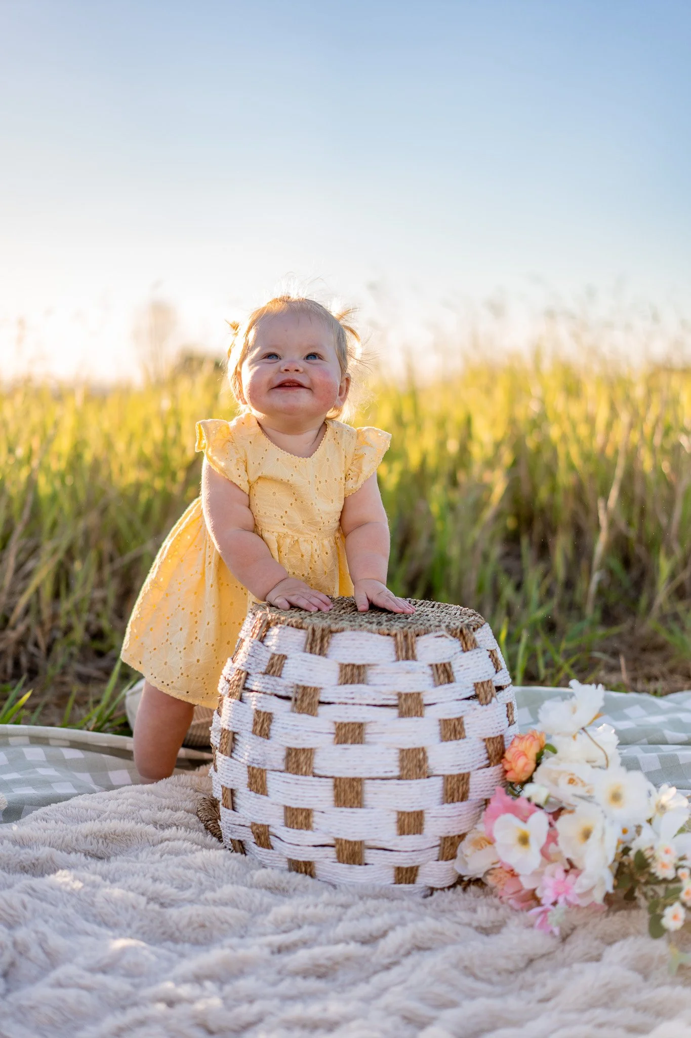 A smiling baby girl in a yellow dress standing outdoors in a field of tall grass, with a white and brown woven basket in front of her, surrounded by flowers, during late afternoon or early evening sunlight.