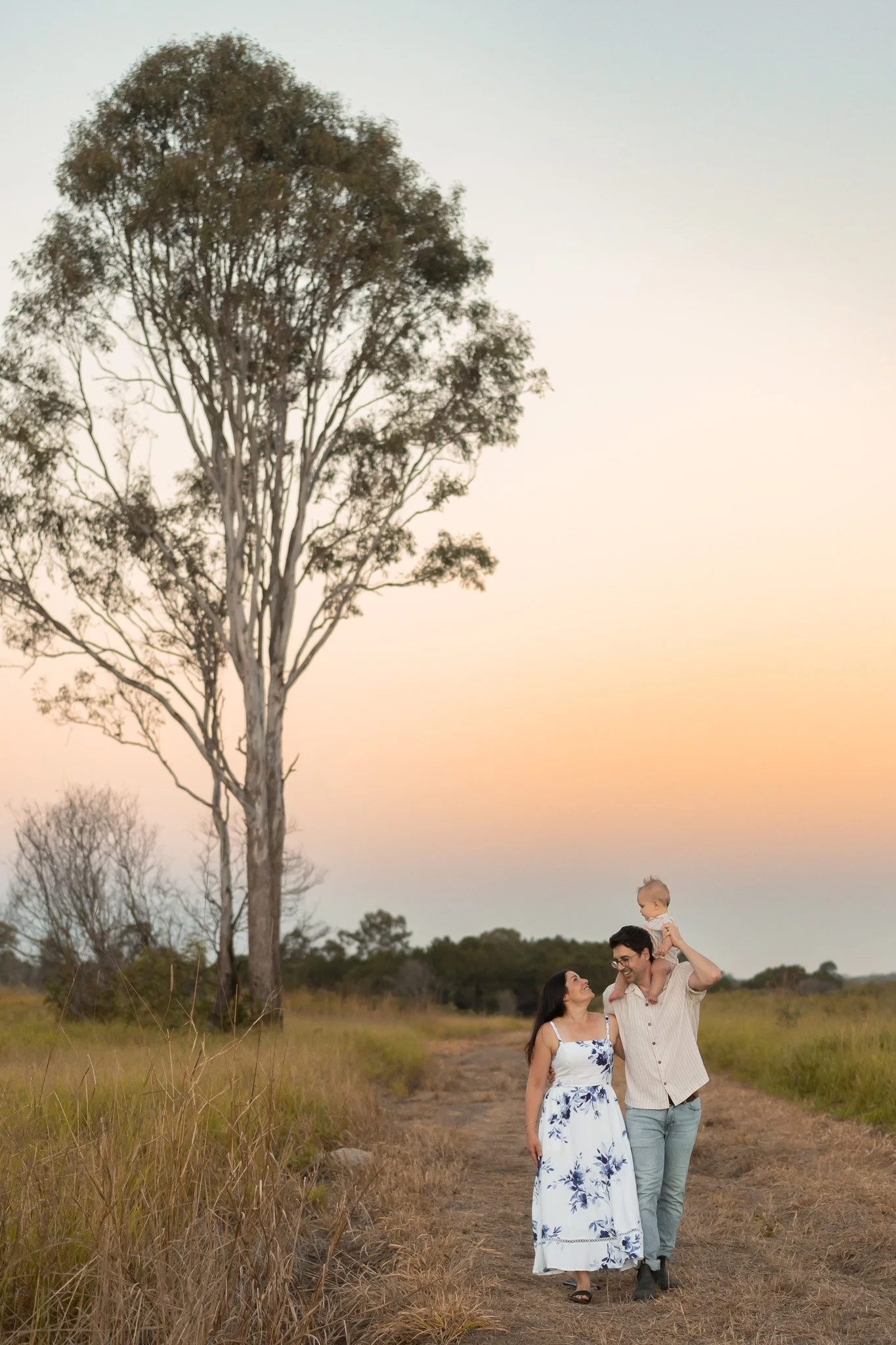 A family of three walking on a dirt path in a grassy field during sunset. The man is carrying a young child on his shoulders, and the woman is walking beside them, smiling. There is a tall tree with sparse leaves behind them, and the sky is a soft gradient of pink, orange, and blue.