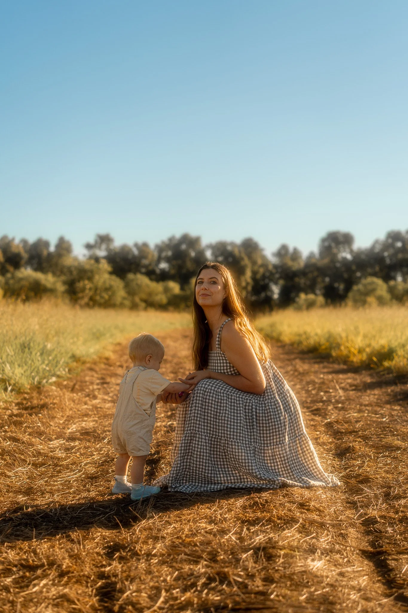A woman and two children in a field on a sunny day. The woman is sitting on the ground wearing a checkered dress, and the children are standing, one holding the woman's hand. The background features trees and a clear blue sky.