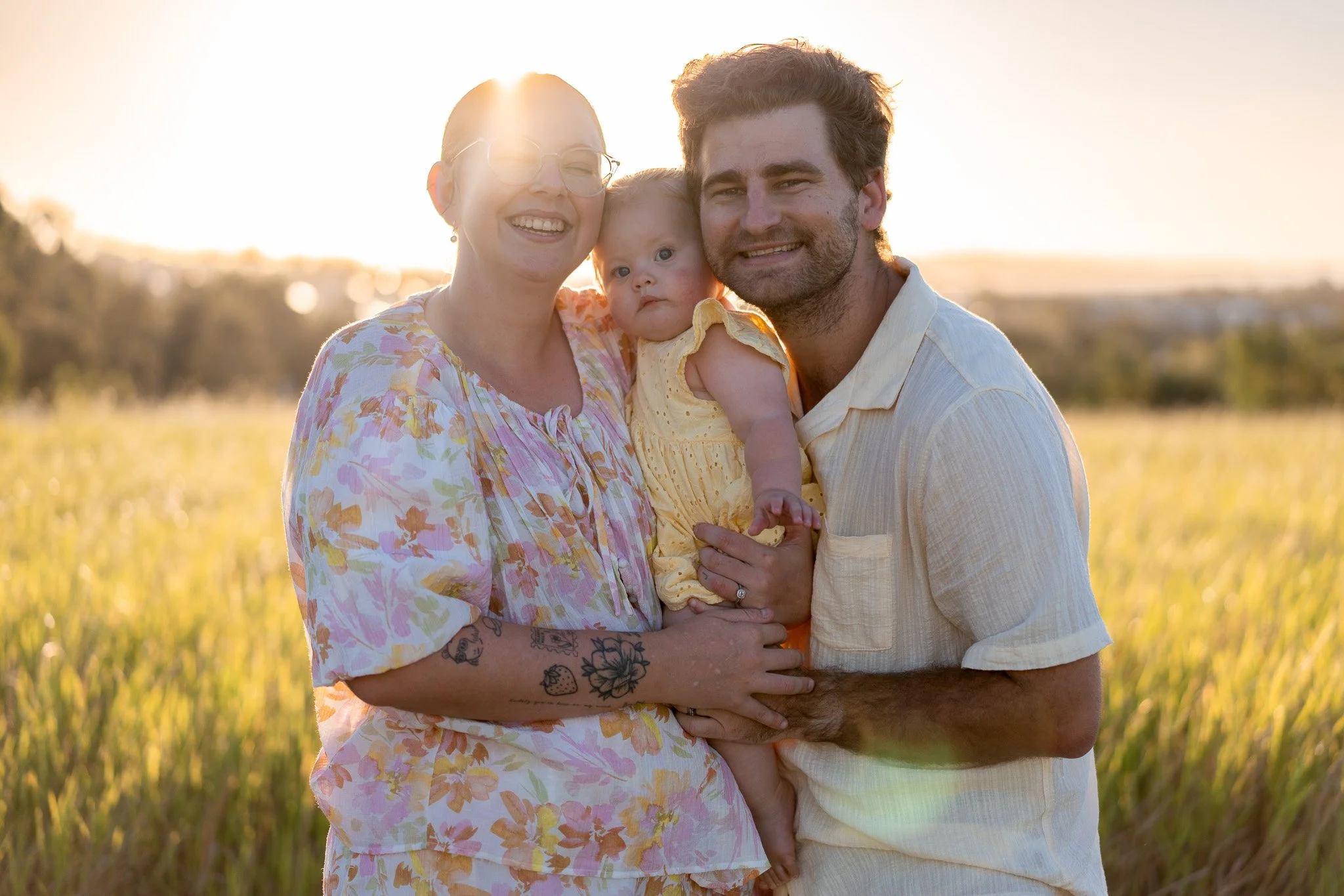 A happy family of three, a woman, a man, and a baby girl, standing outdoors in a field during sunset, smiling at the camera.