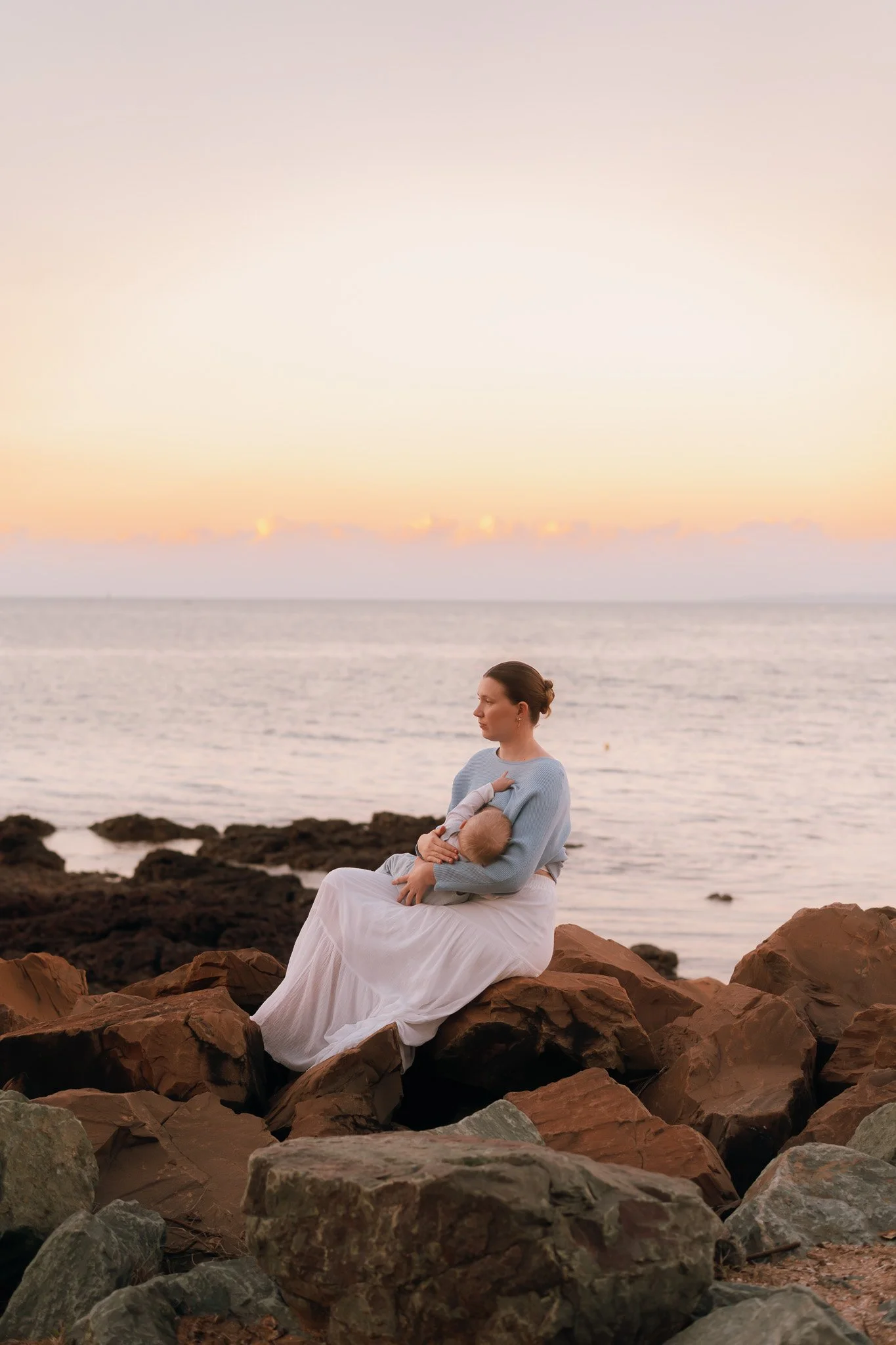 Woman in white dress sitting on rocks by the ocean, breastfeeding her baby during sunset.