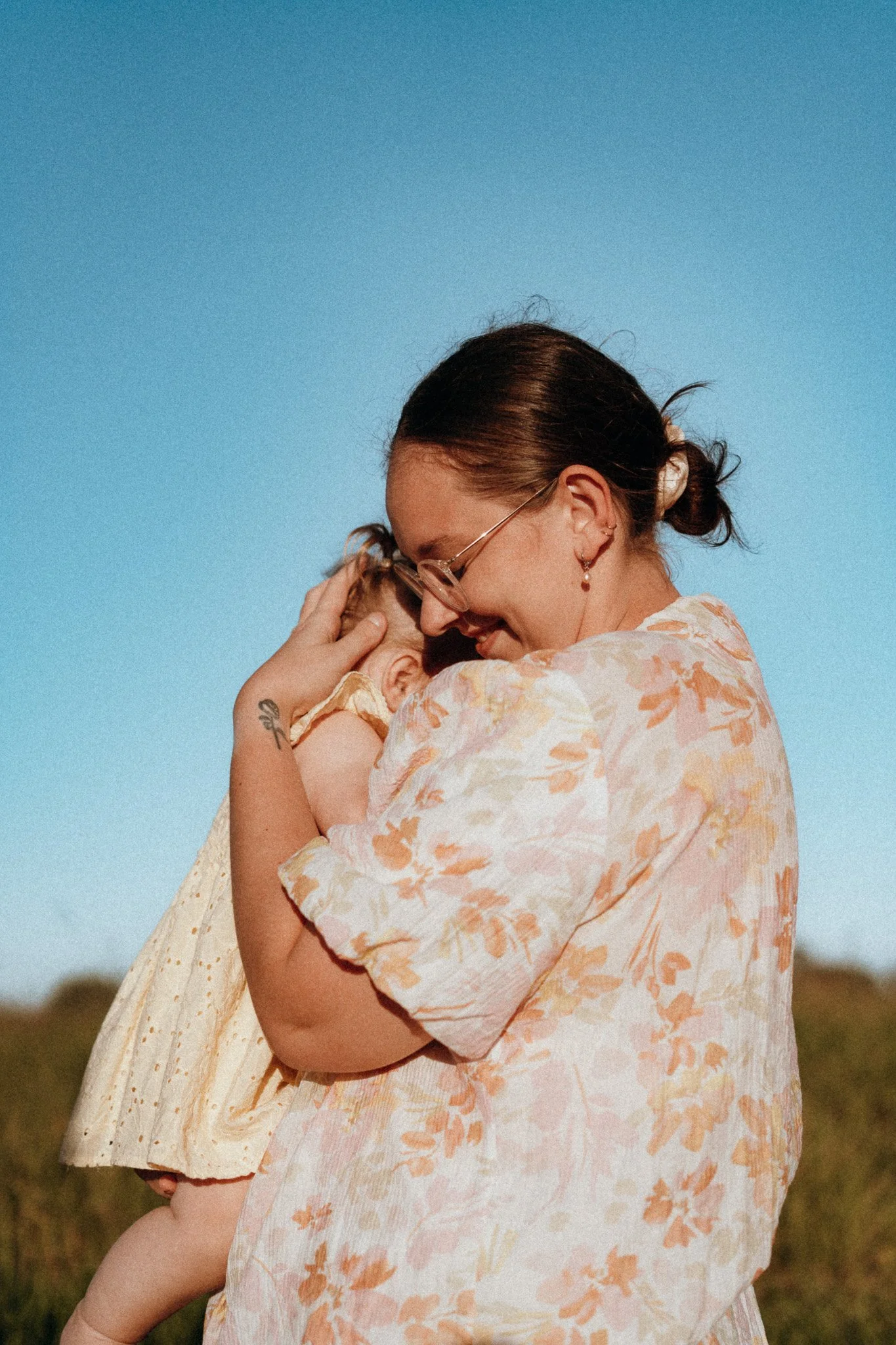 A woman with dark hair tied in a bun, wearing glasses and floral earrings, embraces a young child, both dressed in light-colored clothing with floral patterns. They are outdoors under a clear blue sky.