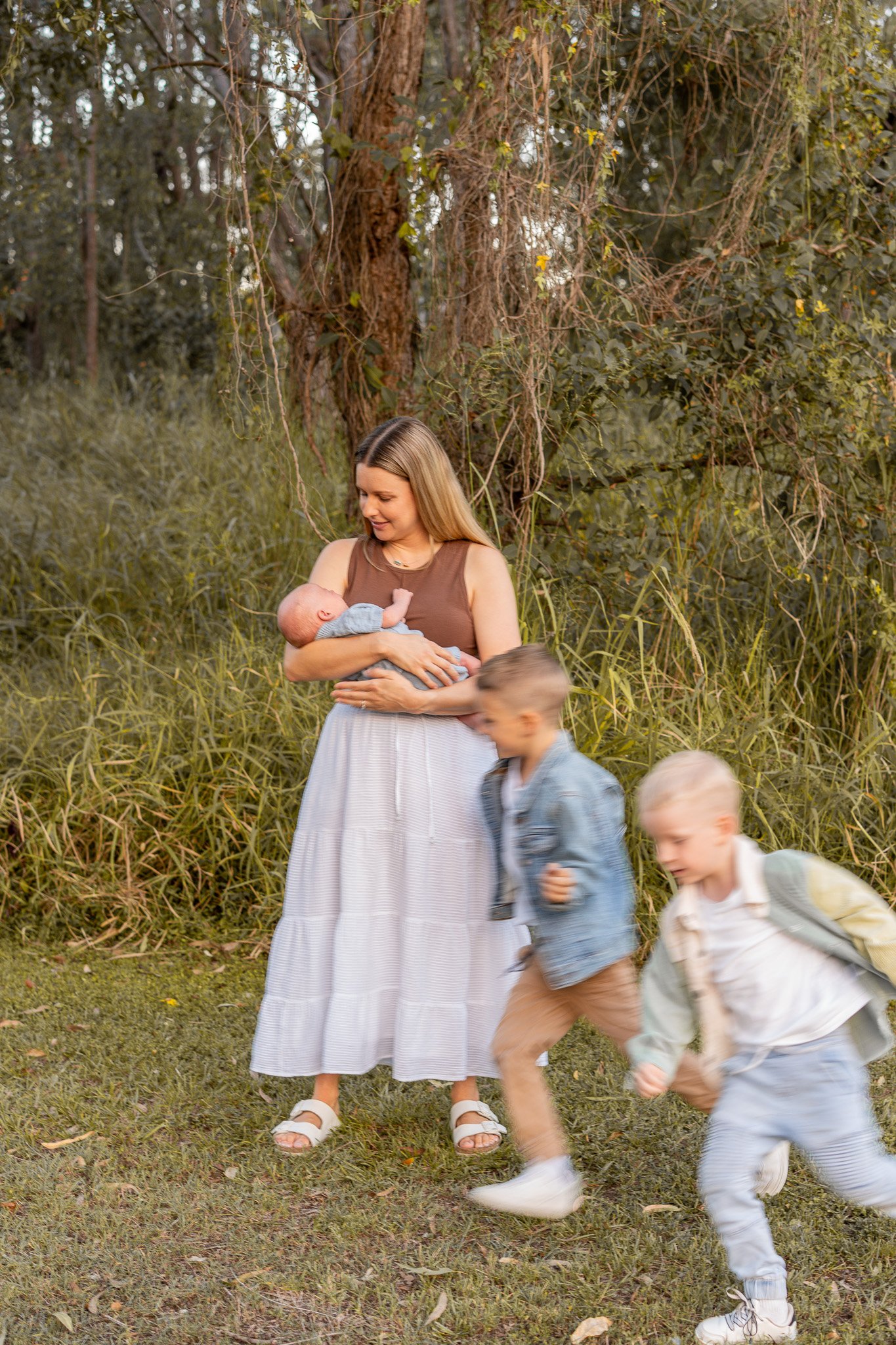 Woman holding a baby outdoors with children running nearby in a grassy area surrounded by trees and foliage.