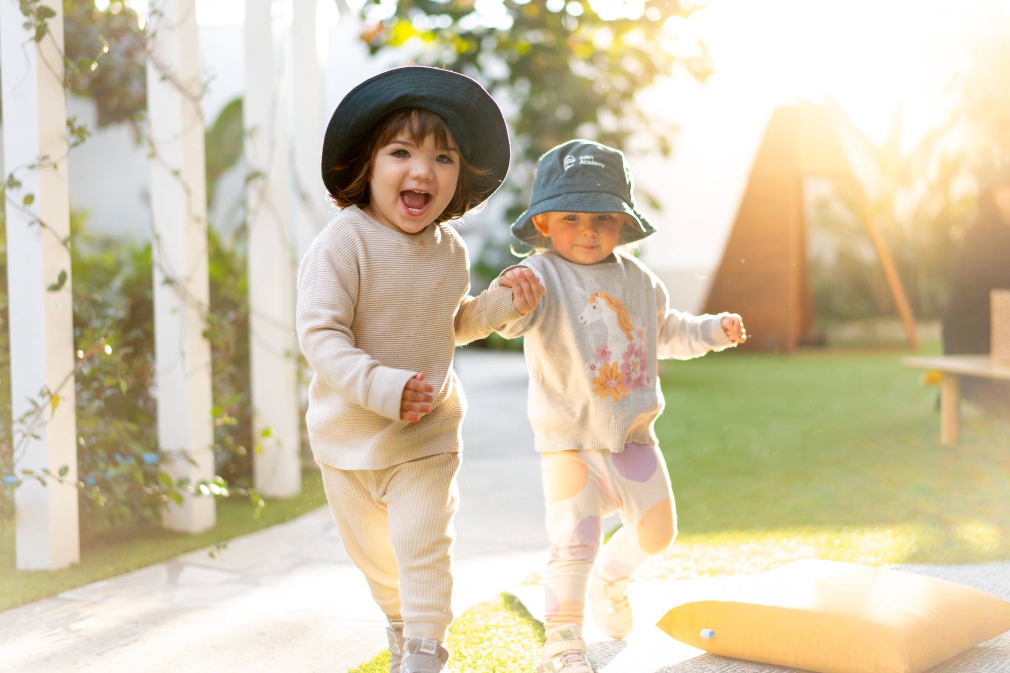 Two young girls wearing sun hats and casual clothing, playing outdoors in sunlight, near a white fence and trees.
