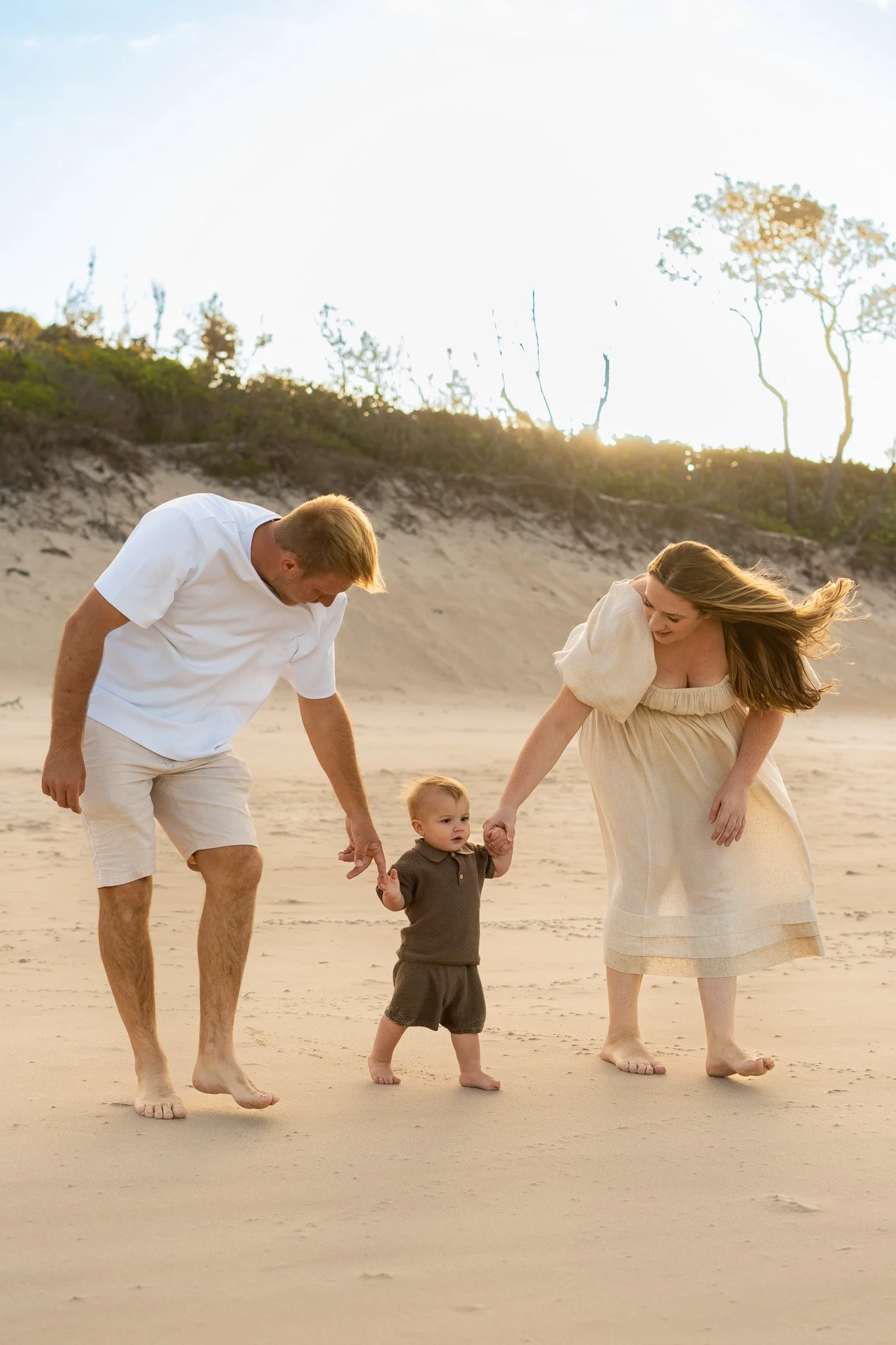 A family of three, including a man, a woman, and a young child, holding hands and walking barefoot on a beach during sunset, with sand dunes and sparse trees in the background.