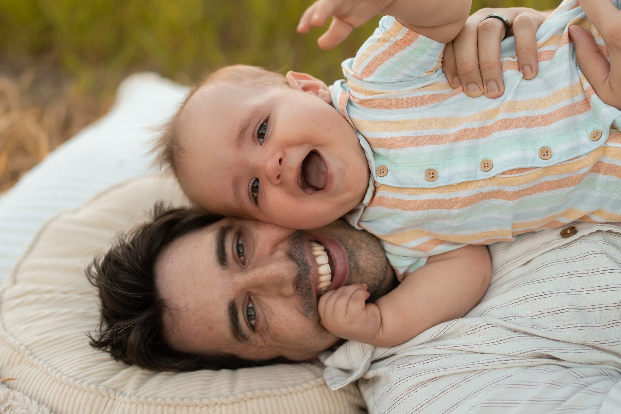 A man lying on a bed with a young child, possibly his son, on top of him. The child is smiling and has his mouth open, wearing a striped shirt. The man is also smiling, showing his teeth, and the background is blurred outdoors.