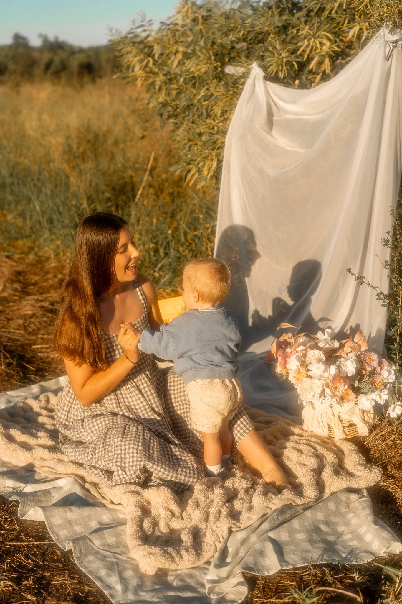 A woman and a young boy sitting on a blanket outdoors with a white fabric backdrop, surrounded by flowers, in a field at sunset.