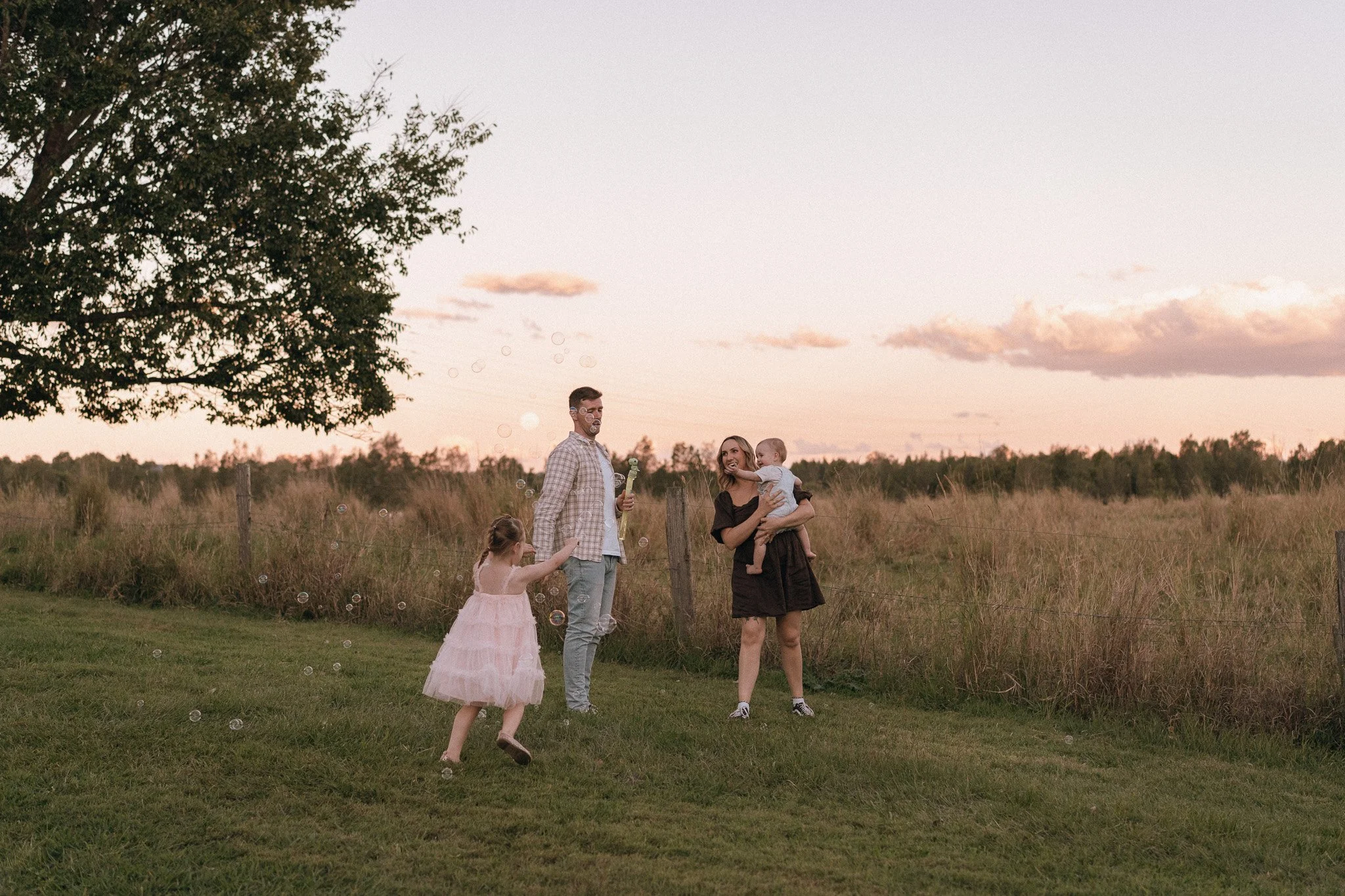 A family of five, including a woman, man, two young children, and an infant, playing and blowing bubbles in a grassy field during sunset with a large tree on the left and clouds in the sky.