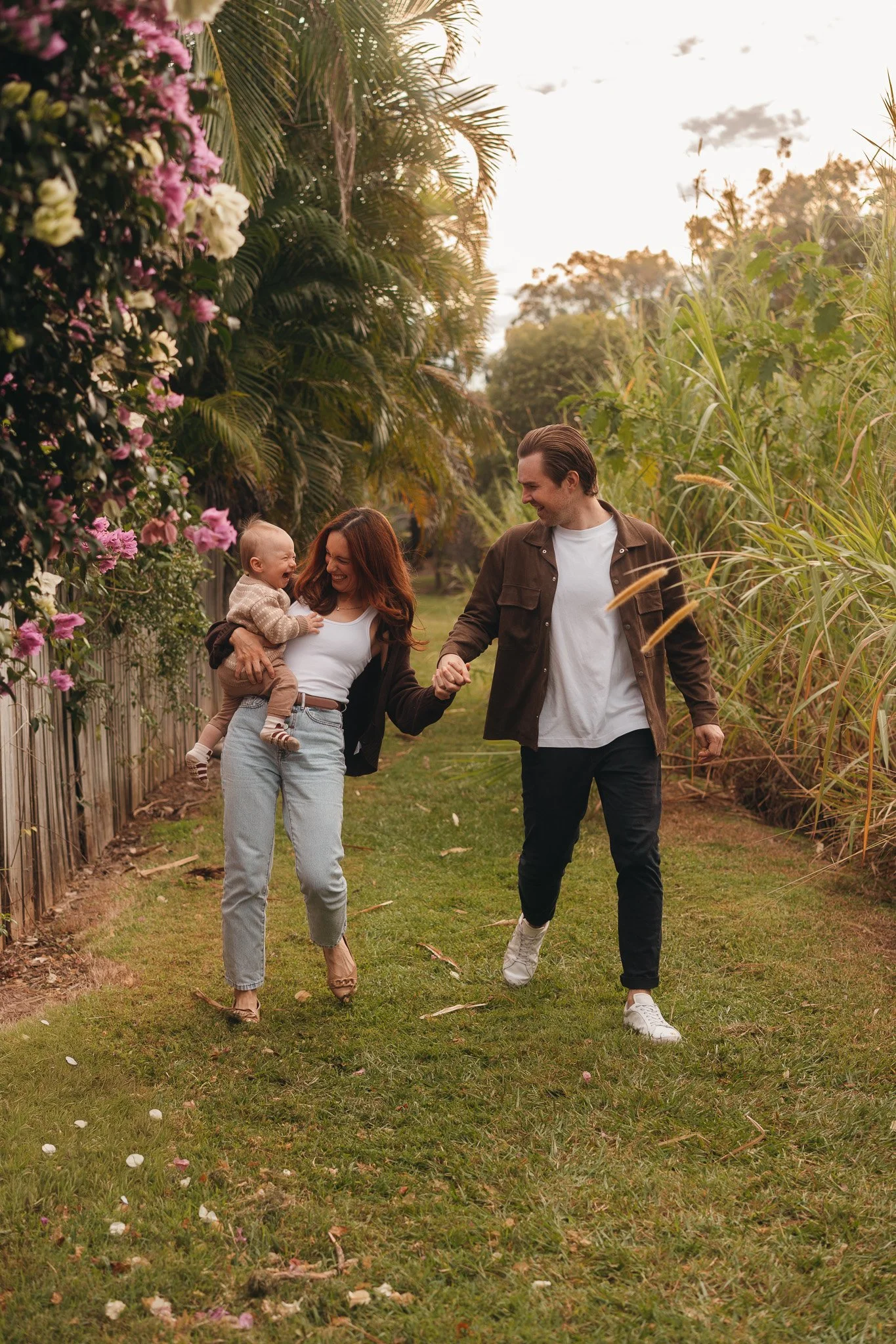 A family of three walking through a lush garden at sunset, laughing and holding hands, with flowers and tall grass surrounding them.