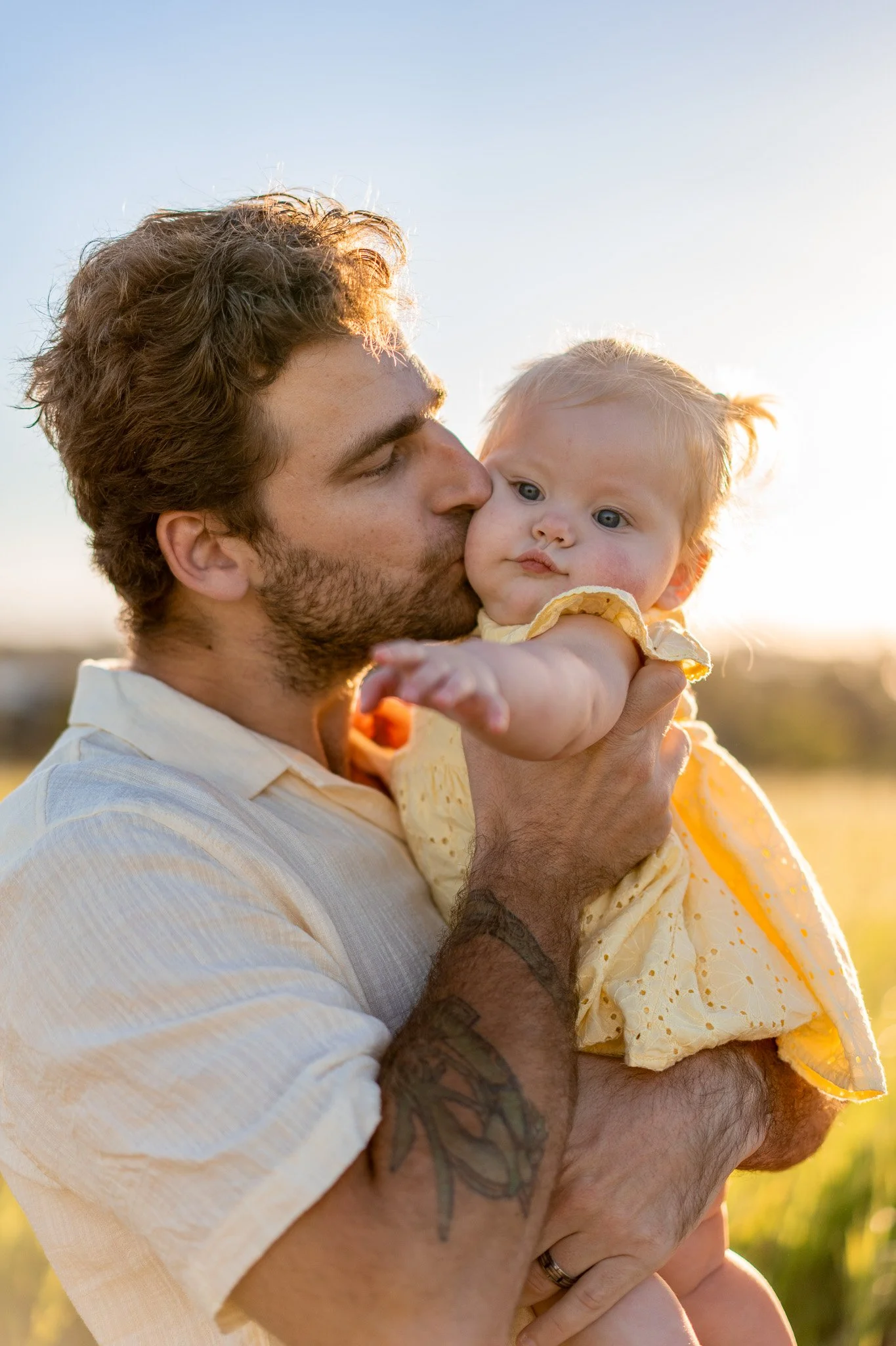 A man with a beard and tattoos holding a young red-haired girl in a yellow dress, kissing her on the cheek outdoors during sunset.