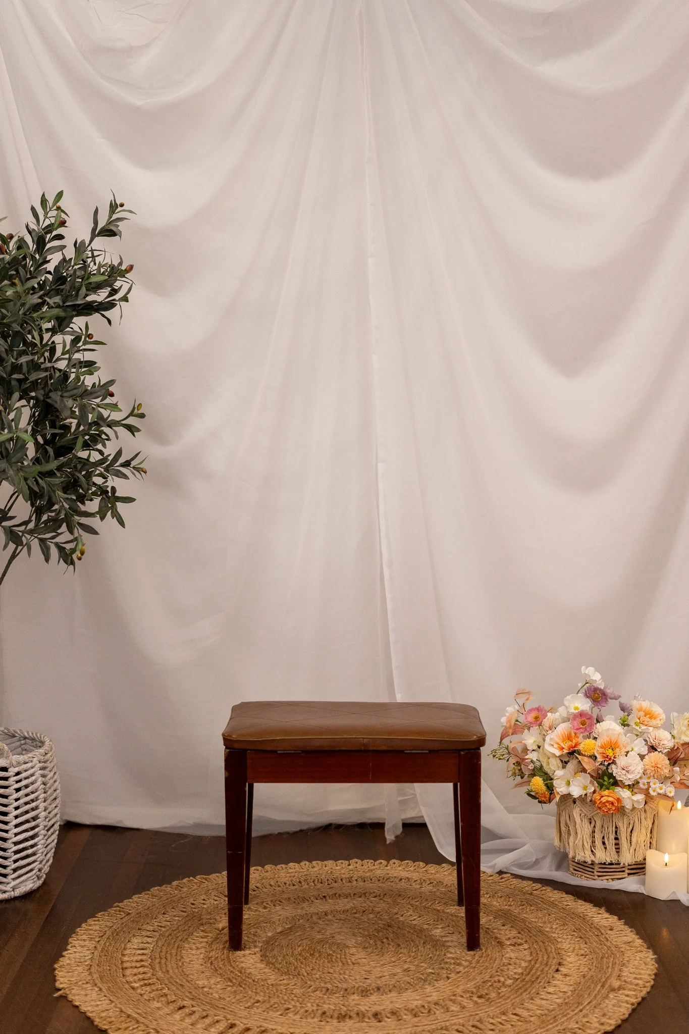 A small wooden table on a round woven rug, with a floral arrangement to the right and candles in front. Behind it, white draped fabric serves as a backdrop, and a green leafy plant is on the left side.