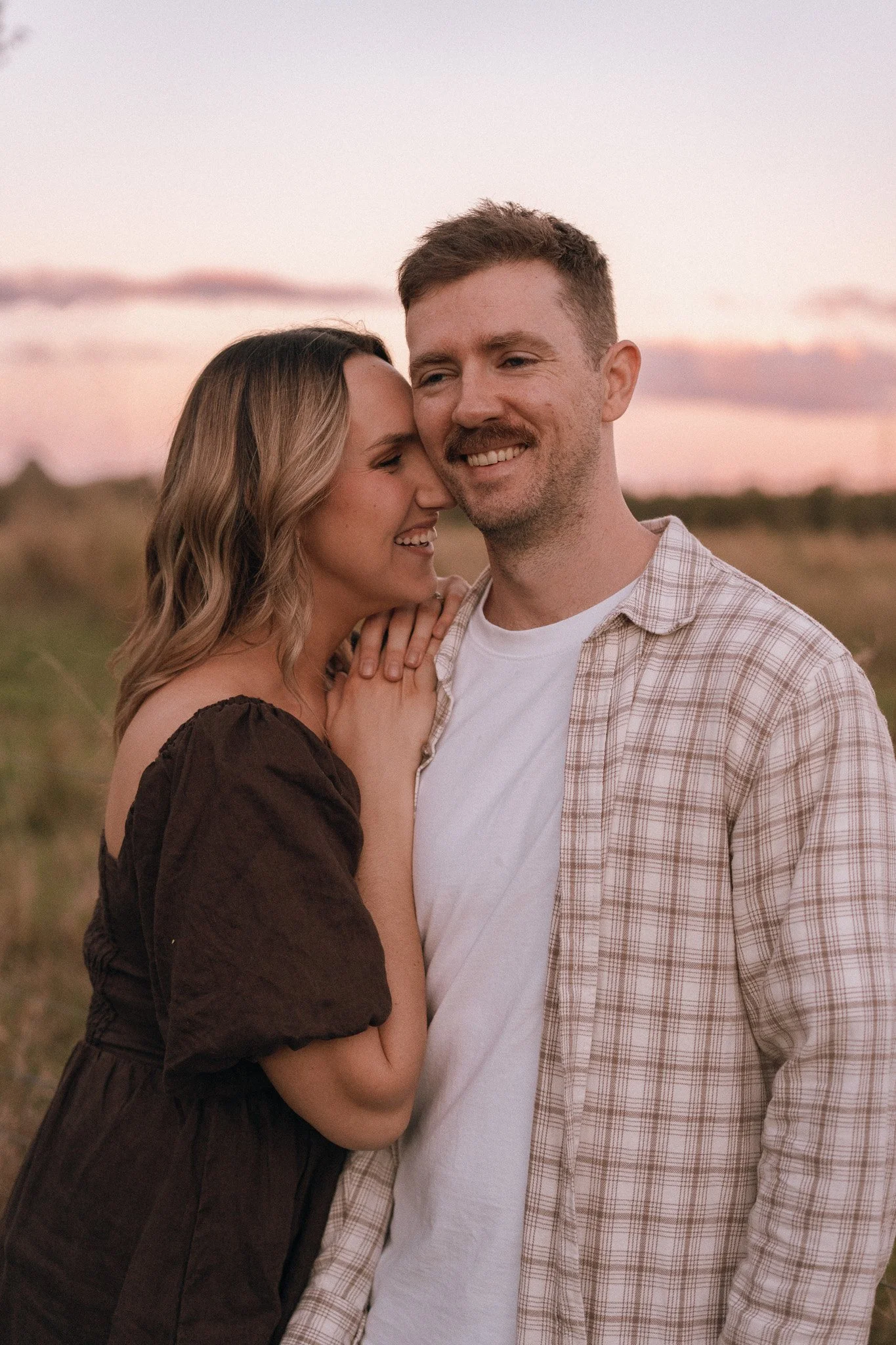 A couple embraces outdoors during sunset, smiling at each other. The woman has blonde hair and is wearing a dark off-shoulder dress, the man has short light brown hair and is wearing a white T-shirt with a plaid shirt over it.