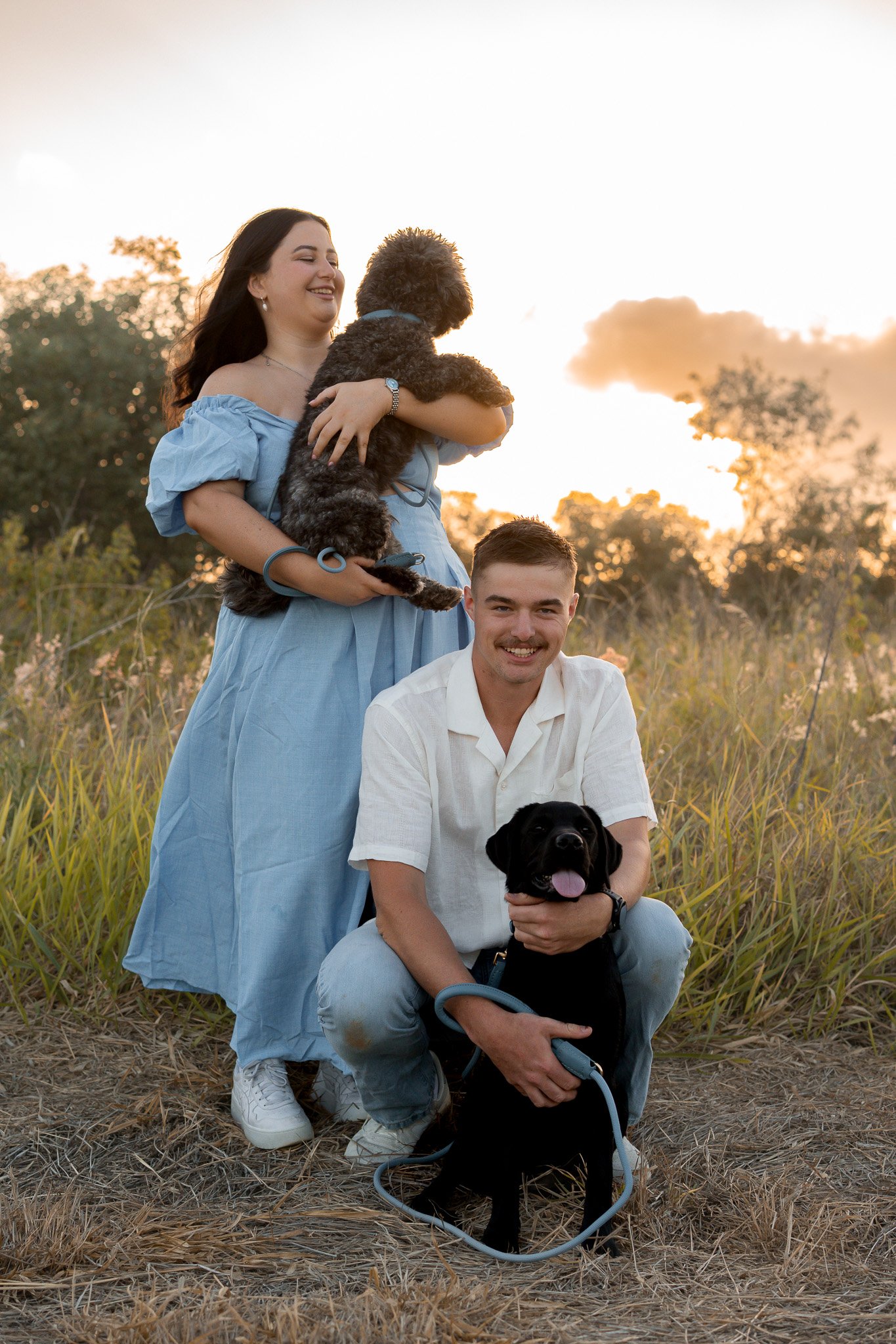 A woman holding a black and white dog, with a man crouching in front of them holding another black dog, in a field at sunset.