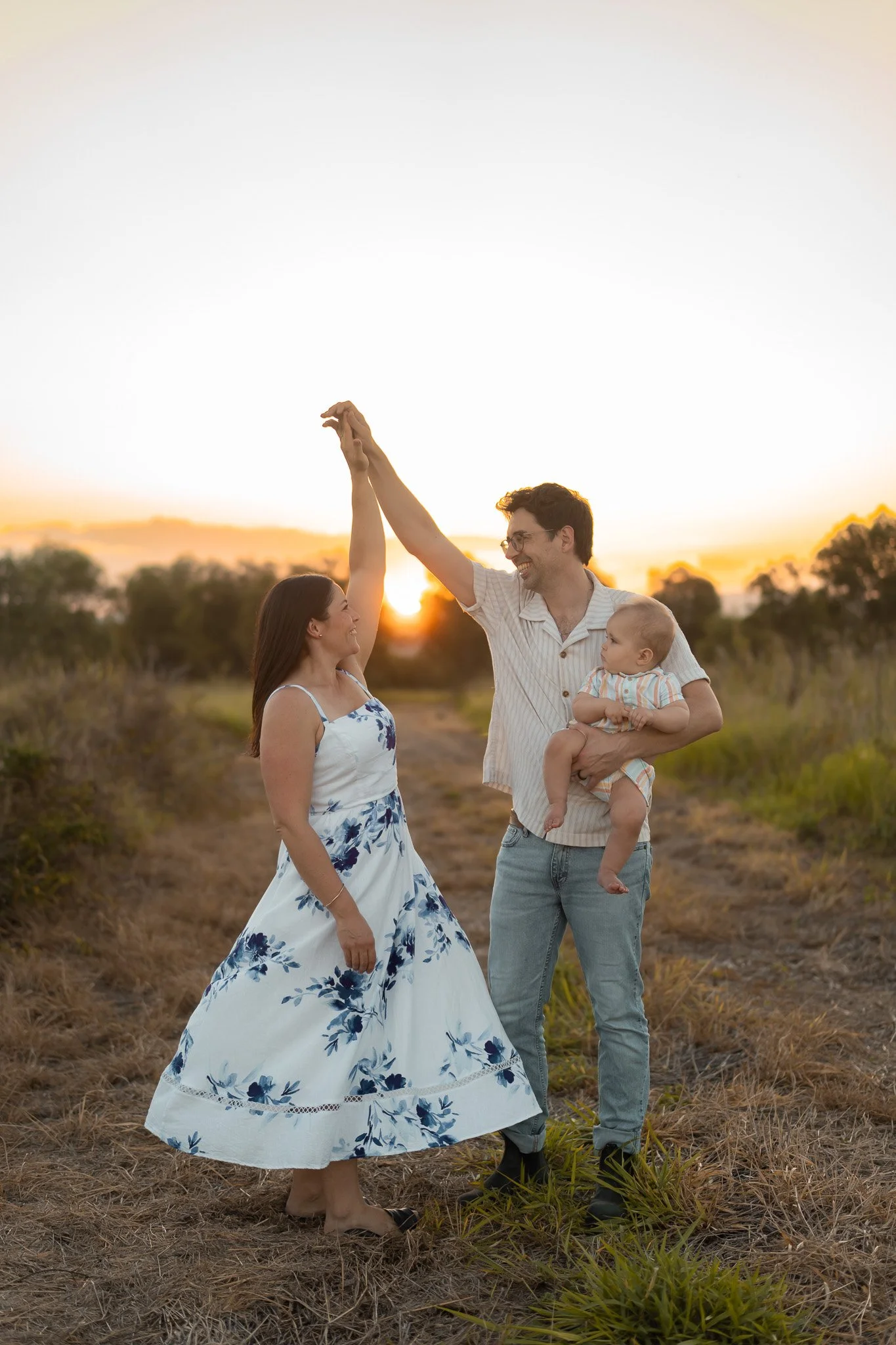 A family of three, a man, a woman, and a baby, standing in a field during sunset, with the man and woman holding hands and smiling.