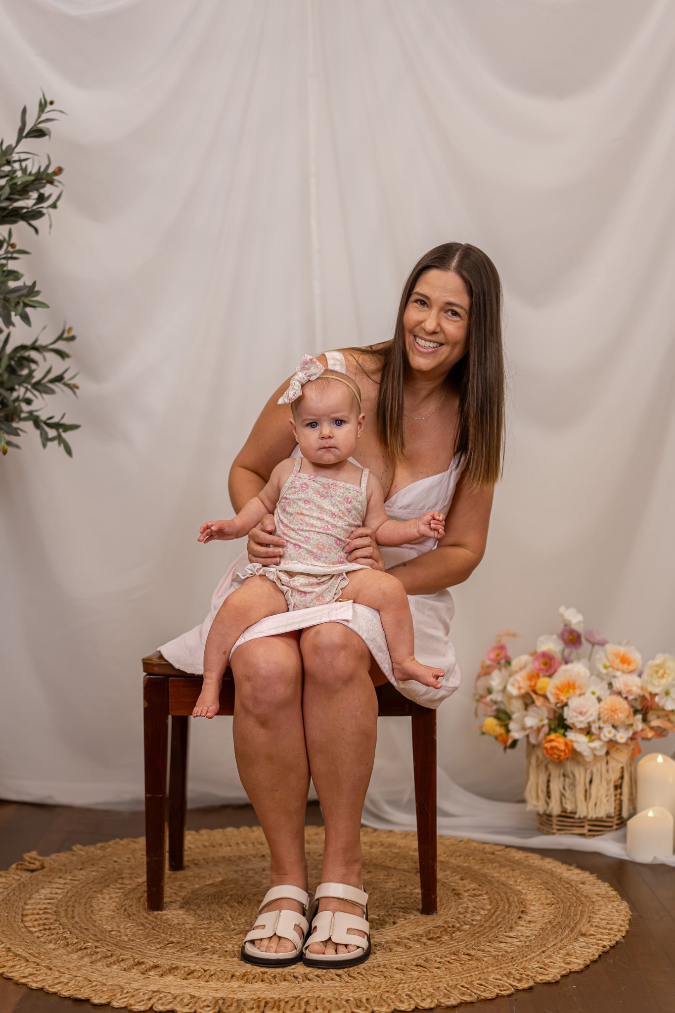 A woman with long brown hair sitting on a wooden stool, holding a young girl on her lap, both smiling at the camera. They are in a decorated room with white drapery background, a basket of flowers, and candles on the floor.