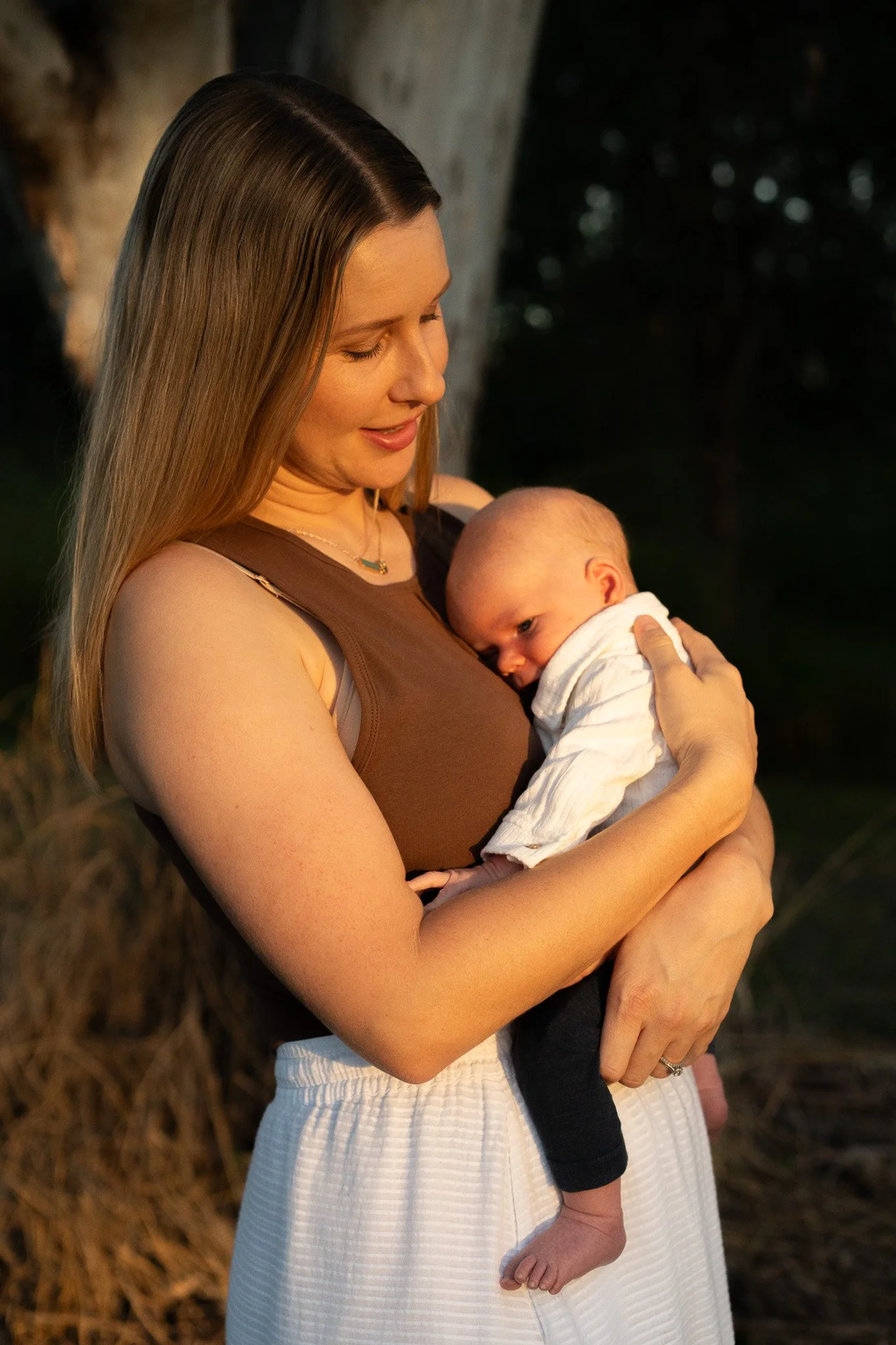A woman holding a baby outdoors during sunset, with a tree and dark forest in the background, both smiling gently.