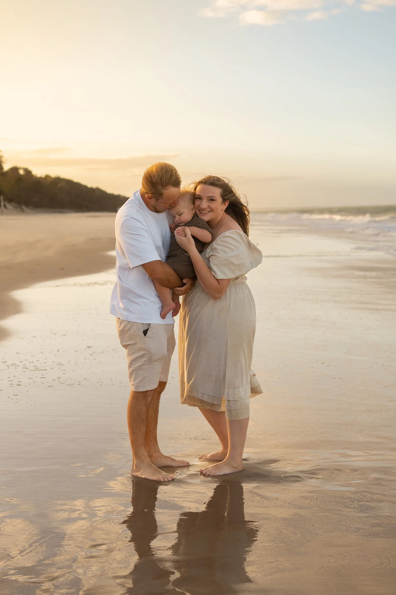 A family of three standing barefoot on the beach at sunset, holding their young child close and smiling at the camera.