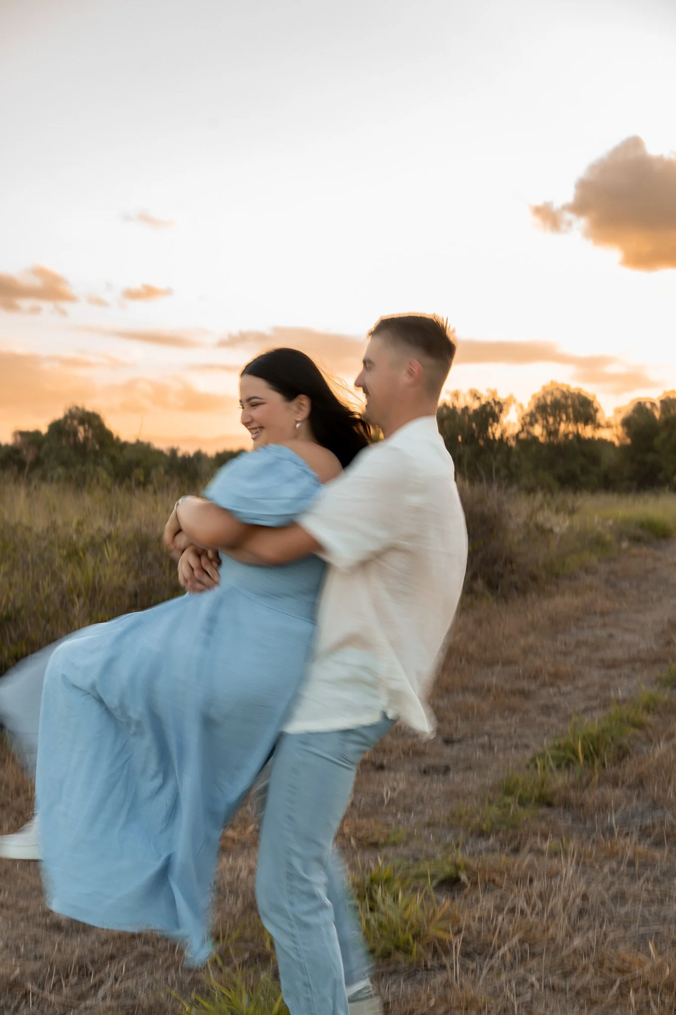 A couple is outdoors during sunset, with the woman in a blue dress being carried and embraced by the man, both smiling and enjoying the moment.