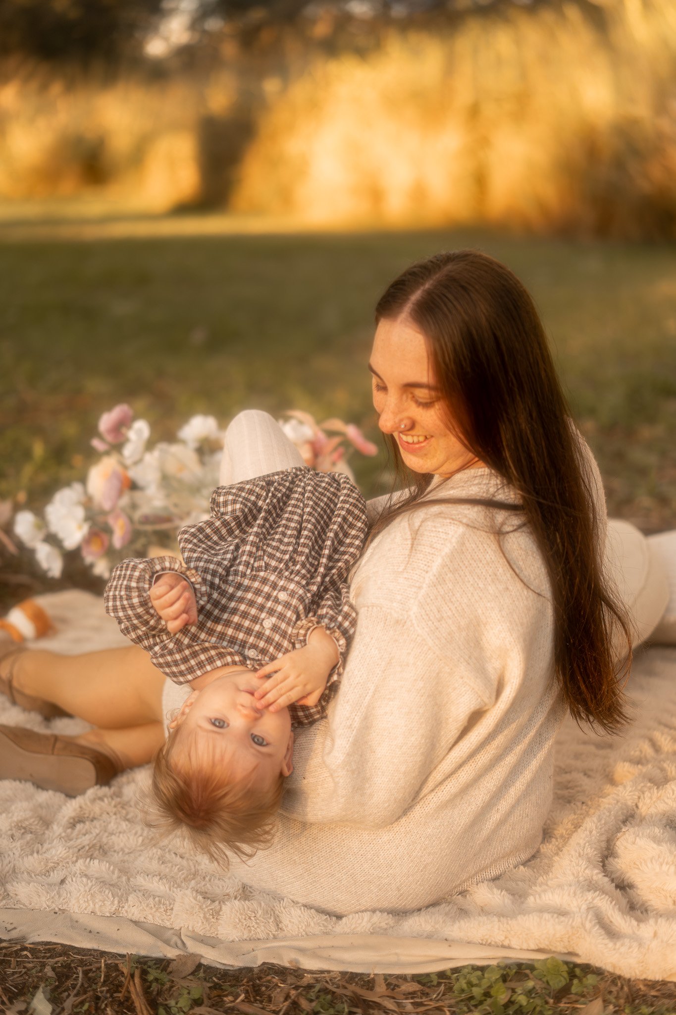 A woman is sitting on a fluffy rug outdoors, holding a young girl upside down. The woman is smiling and has long, brown hair. The girl, with light brown hair, is lying on the rug and looking at the camera, with her head upside down and wearing a plaid dress. There are flowers on the rug and a blurred background of trees with autumn leaves.