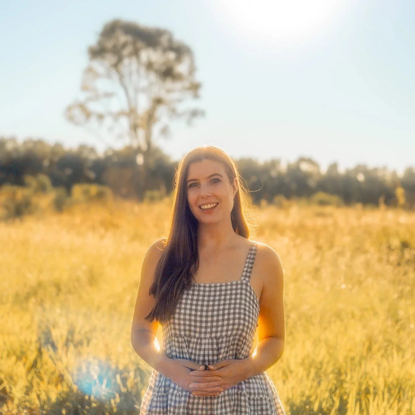 A young woman with long dark hair smiling in a sunlit field during golden hour, wearing a checkered dress.