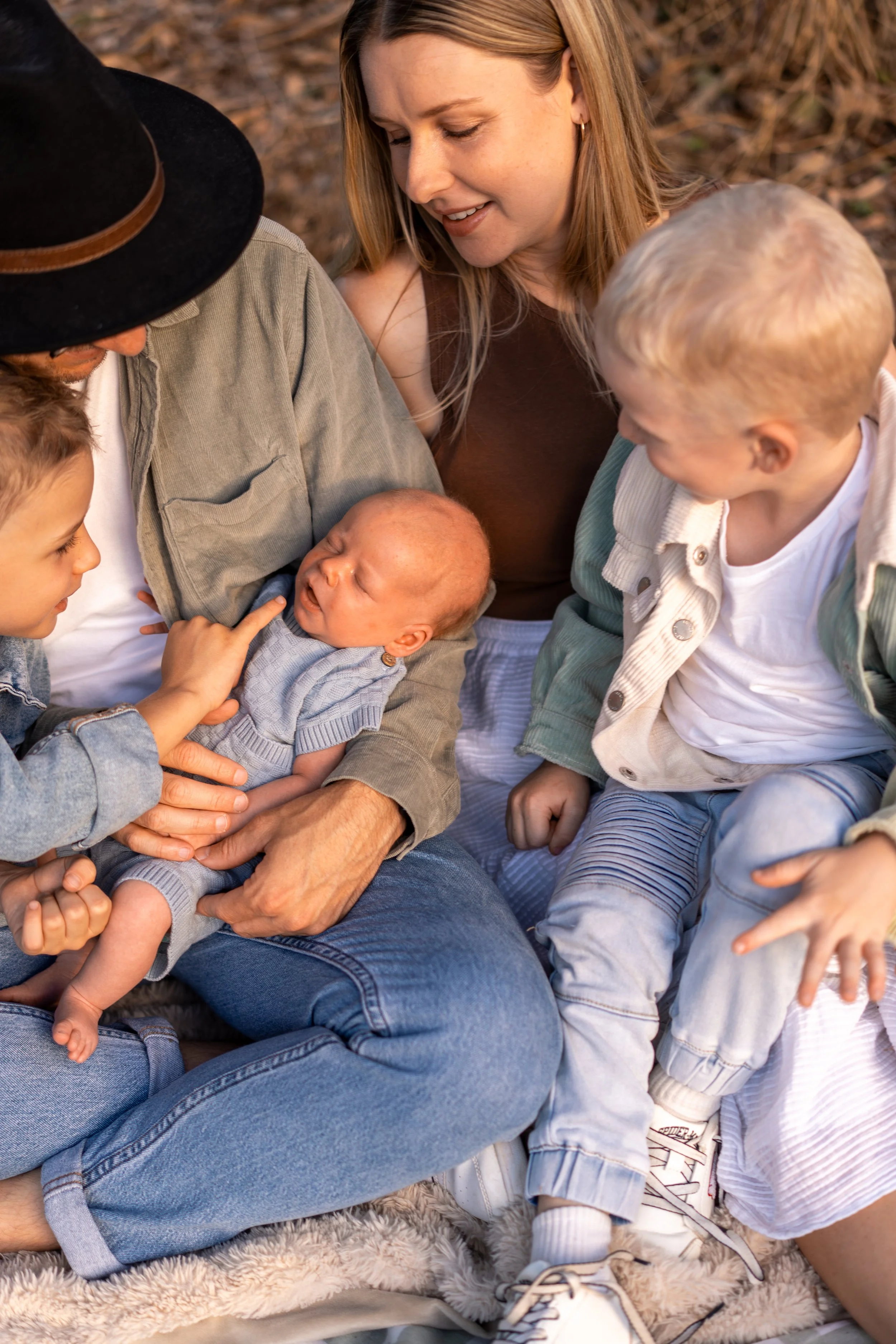 A woman with long blonde hair and light skin, smiling while holding a newborn baby, surrounded by three young children with light skin, outdoors on a sunny day. The children are engaging with the baby, with one touching the baby's face and others nea
