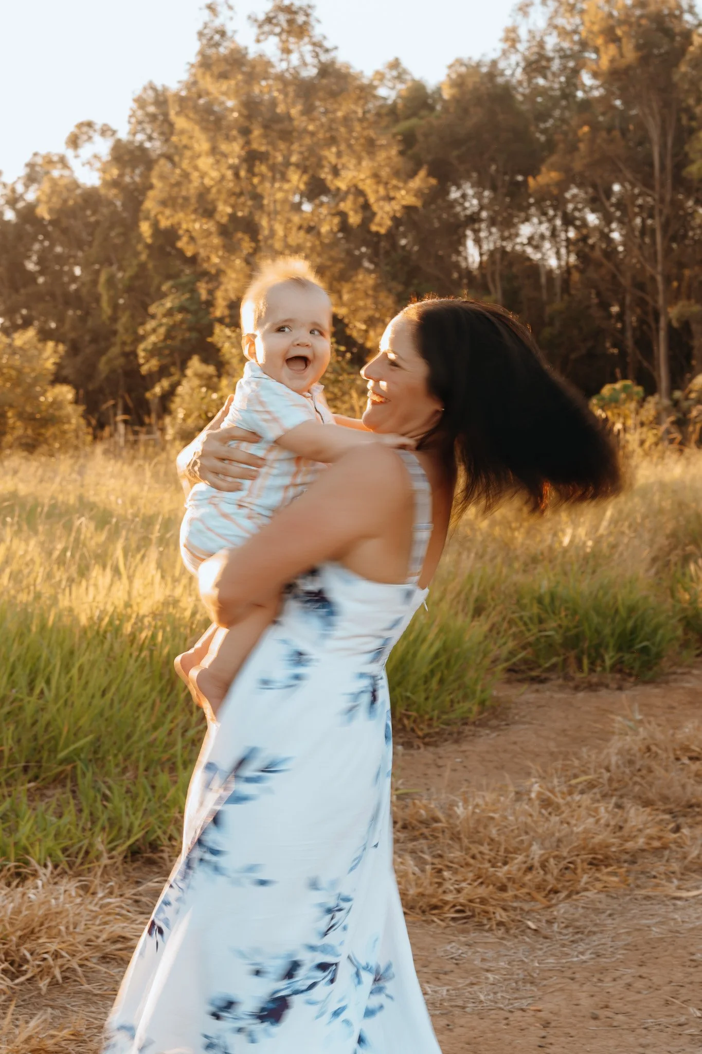 A woman holding a smiling child outdoors during sunset, with trees and tall grass in the background.