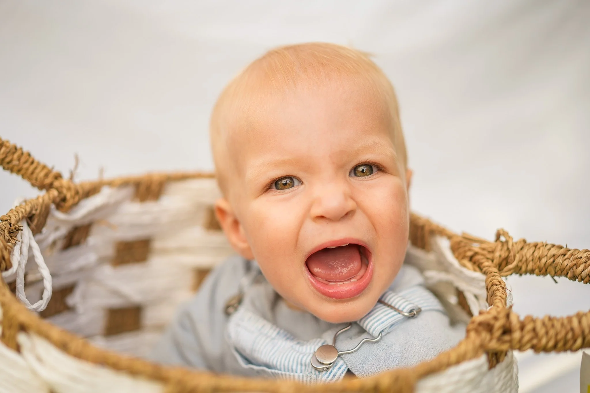 Close-up of a baby with light skin and green eyes, sitting in a woven basket, with mouth open, showing teeth, wearing a striped shirt and a necklace.