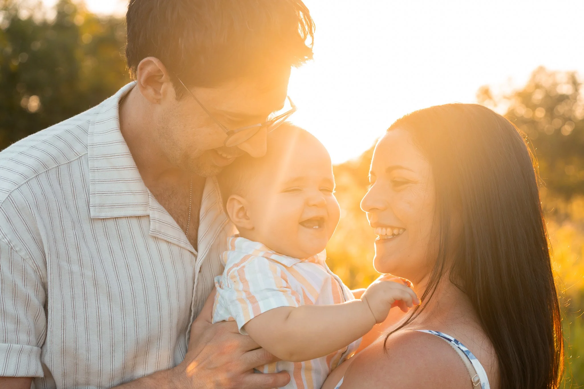 A family of three enjoying sunset, smiling and looking at each other, outdoors with trees in the background.