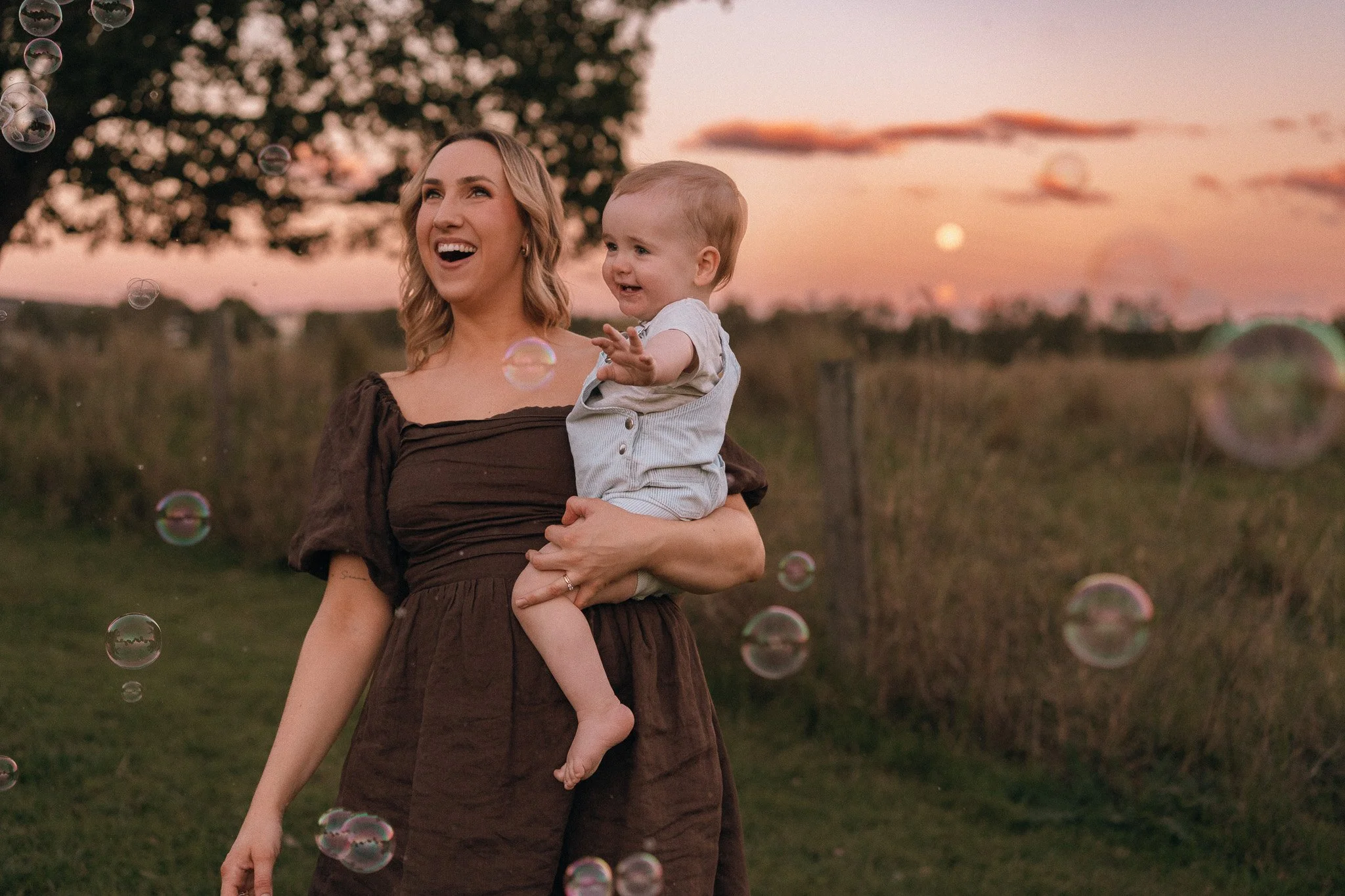 A woman holding a smiling young boy outdoors during sunset, surrounded by floating bubbles, with trees and a pink sky in the background.