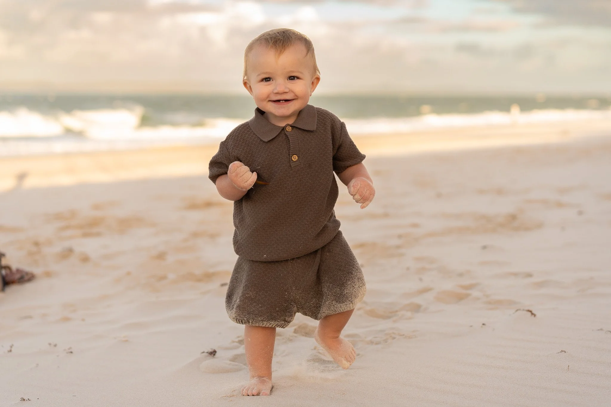 A young boy running on the beach with sand on his hands and feet, wearing a brown shirt and shorts, with the ocean waves in the background.