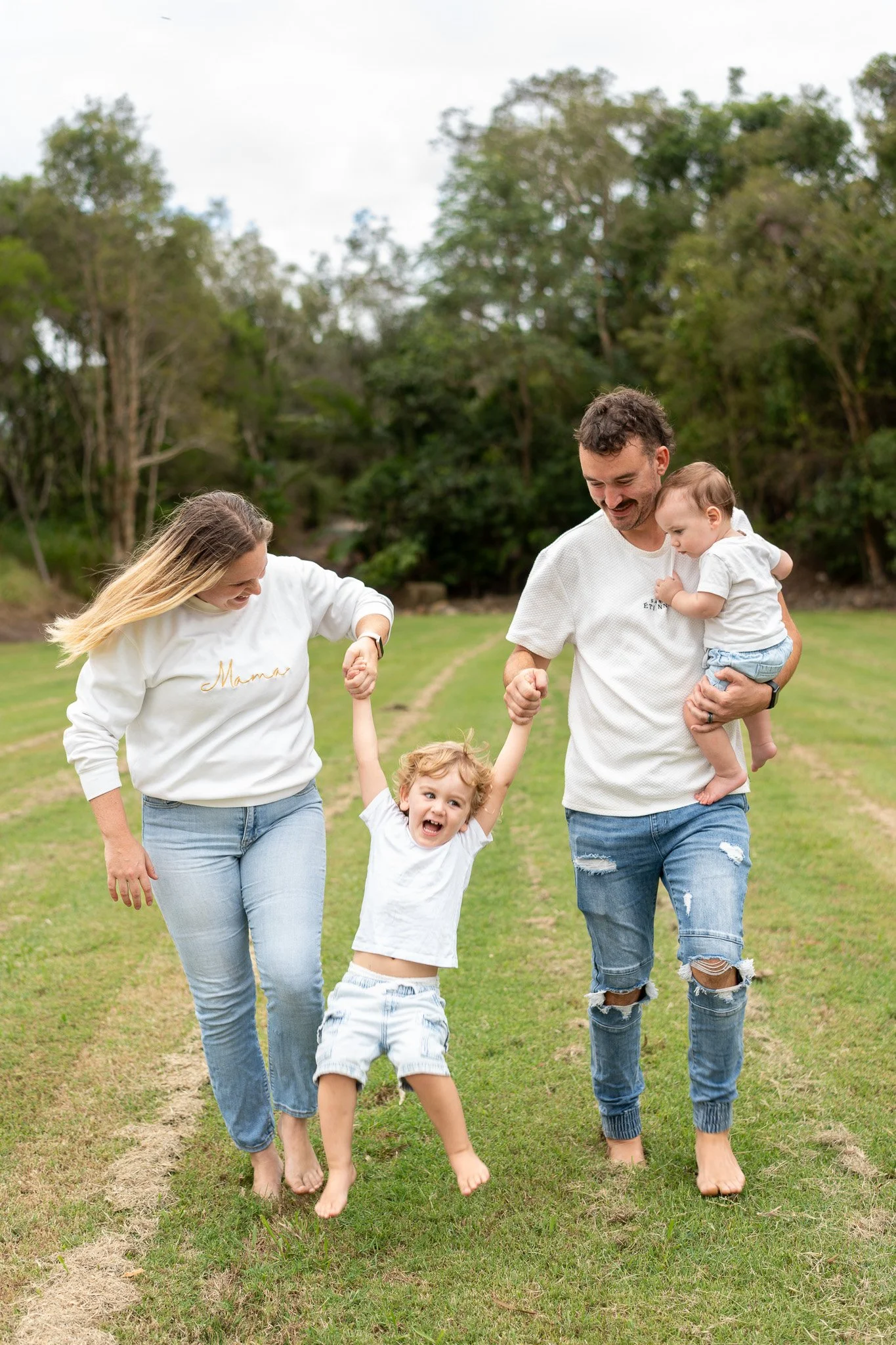A happy family enjoying a day outdoors in the park. The mother and father are holding hands with their two young children, a girl and a boy, who are playing and smiling. The family is dressed casually, and the background features green trees and a grassy field.