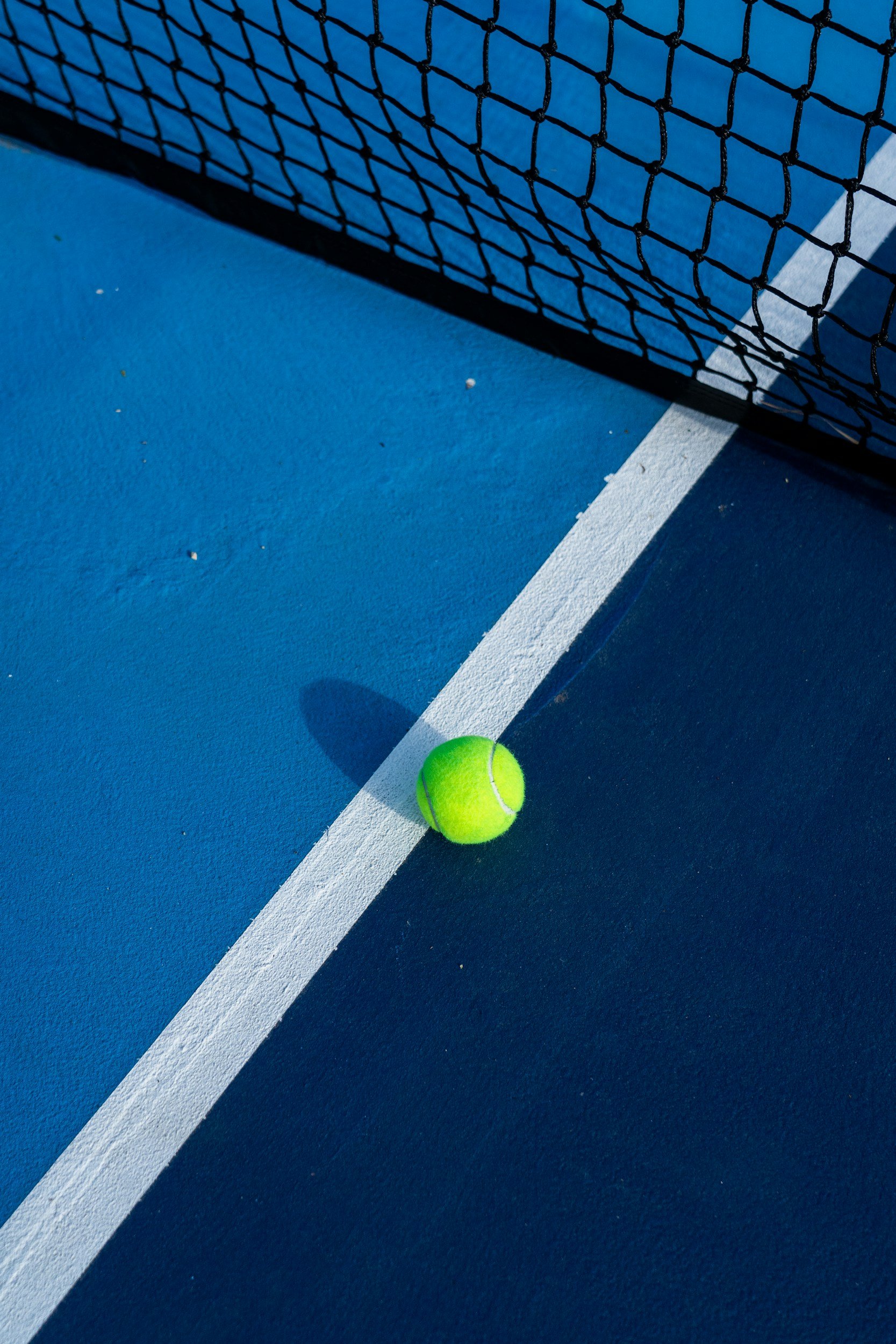 Close-up of a bright green tennis ball on a blue tennis court near a white line, with a black tennis net casting a shadow.