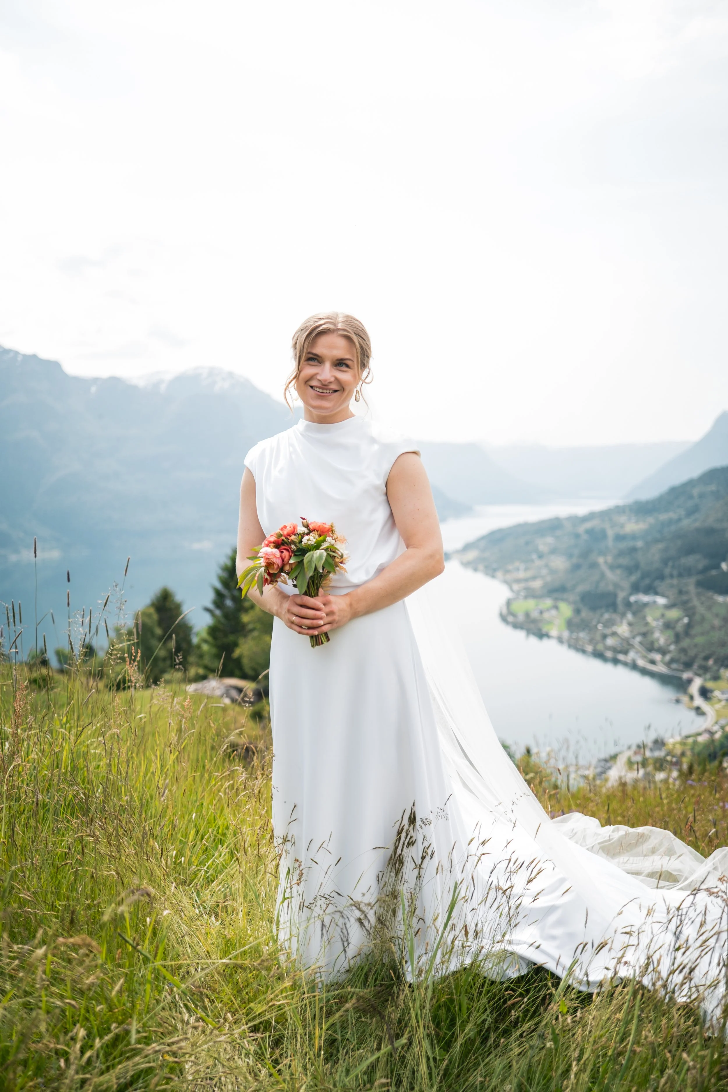 Brud i hvit kjole, markblomster i hånden med sognefjorden i bakgrunnen.