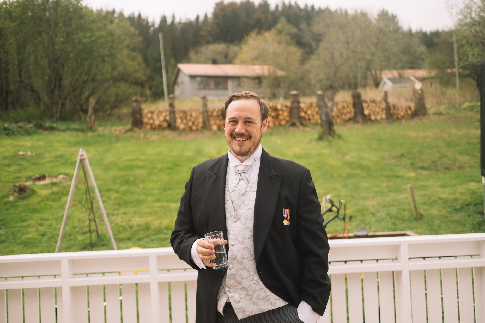En mann i formell dress holder et glass med drikke, smiler, står på veranda med hage og trær i bakgrunnen.