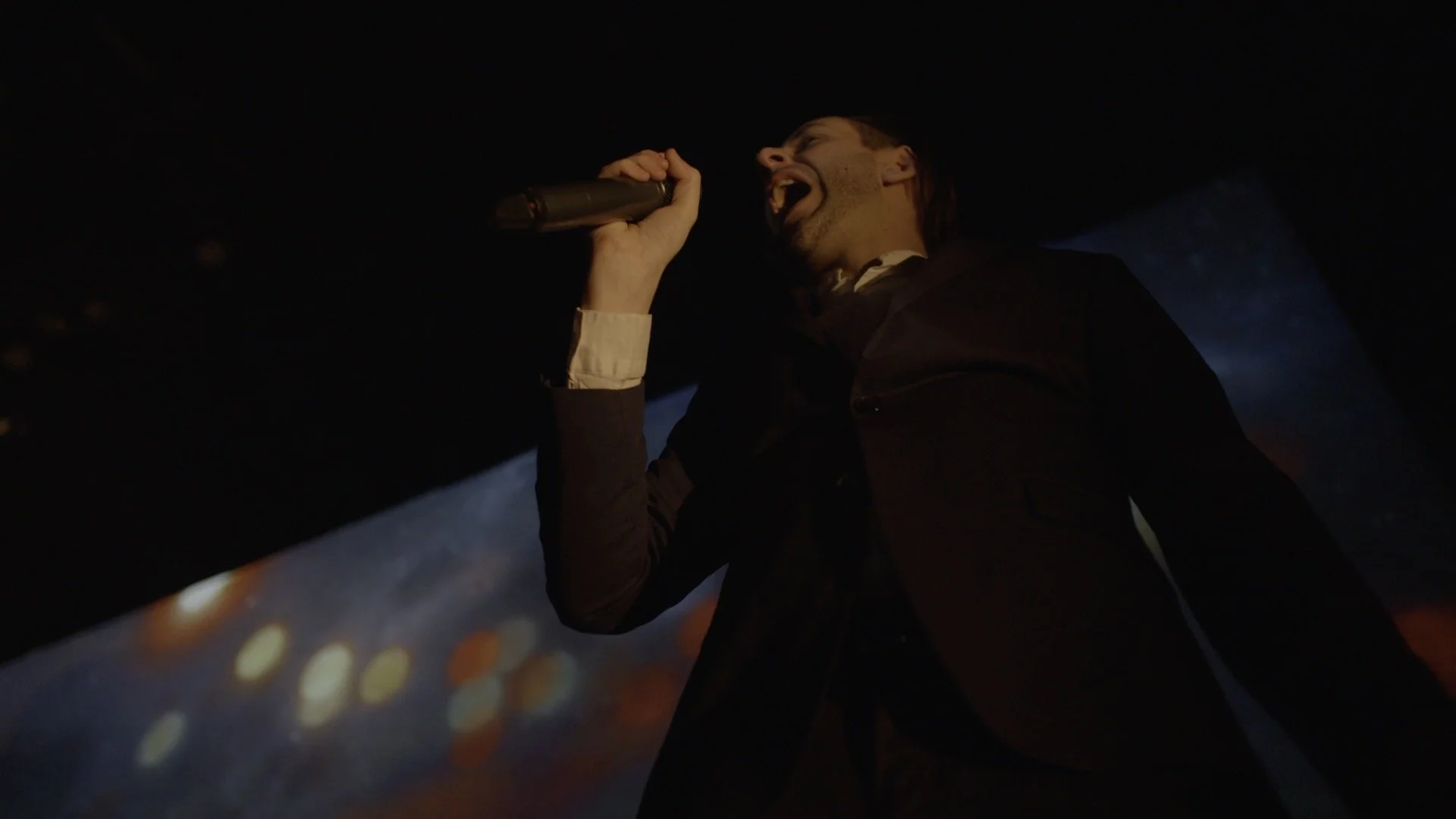 A man in dark clothing singing into a microphone during a performance with a dark background and colorful lights.