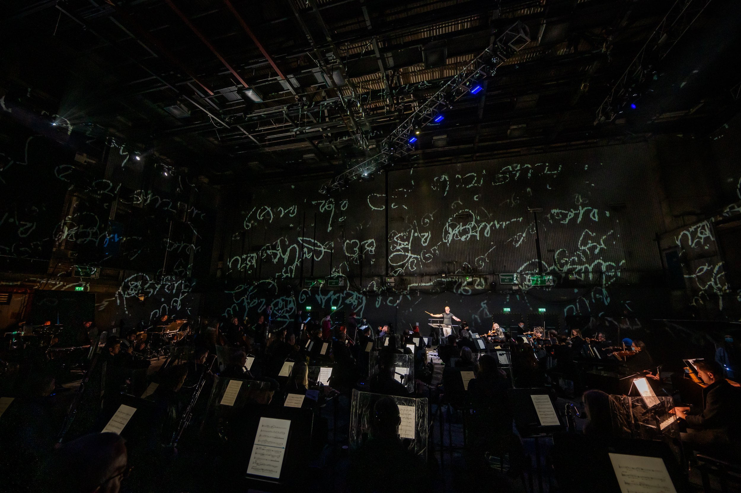 An orchestra performing on stage with a conductor conducting, illuminated by stage lighting and projected writings in the background, in a large dark concert hall.