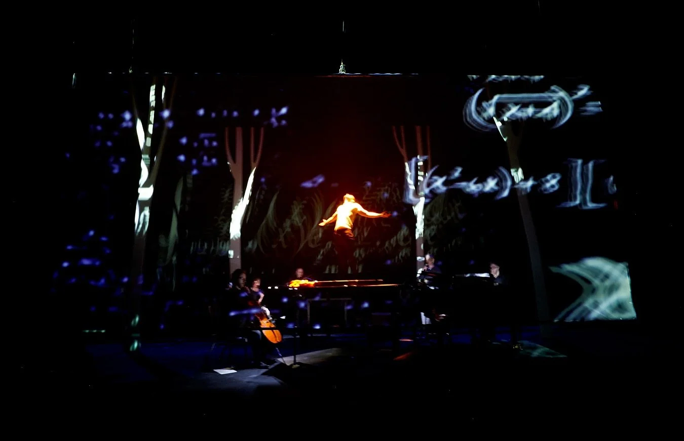 Stage with a performer mid-air, and musicians playing instruments, backdrop with illuminated text and abstract patterns.