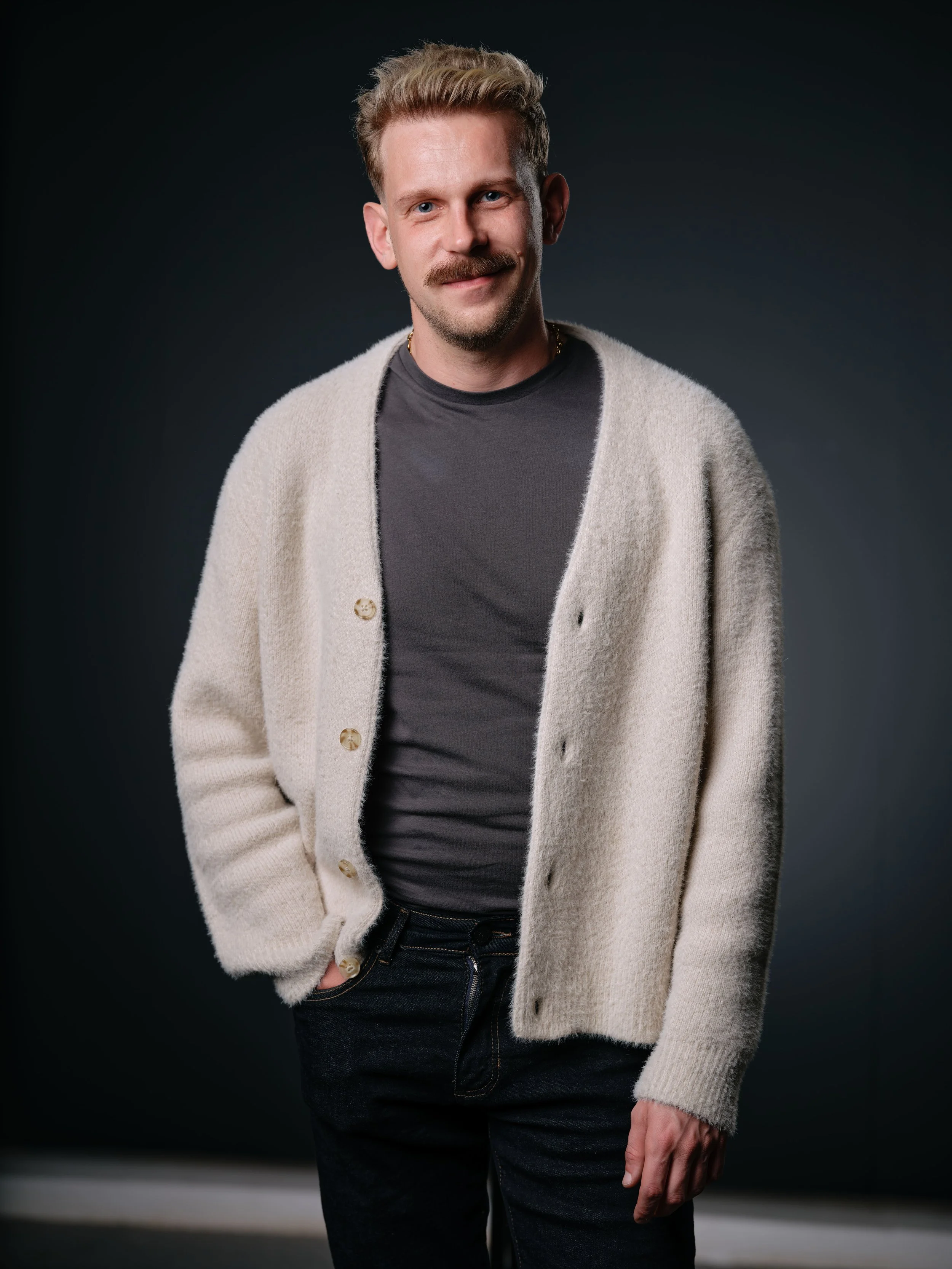 Portrait of a young man with blonde hair and a mustache, wearing a black shirt and a beige cardigan, standing against a dark background.