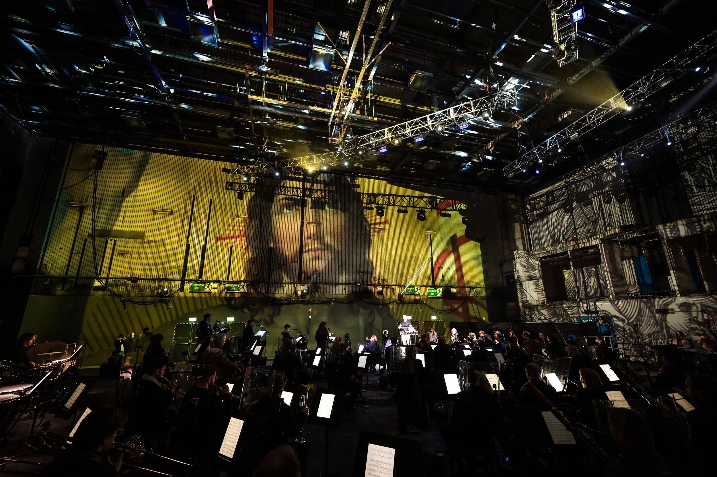 Concert hall with orchestral musicians performing on stage, illuminated with dramatic lighting and reflections, with a large digital screen displaying a woman's face in the background.