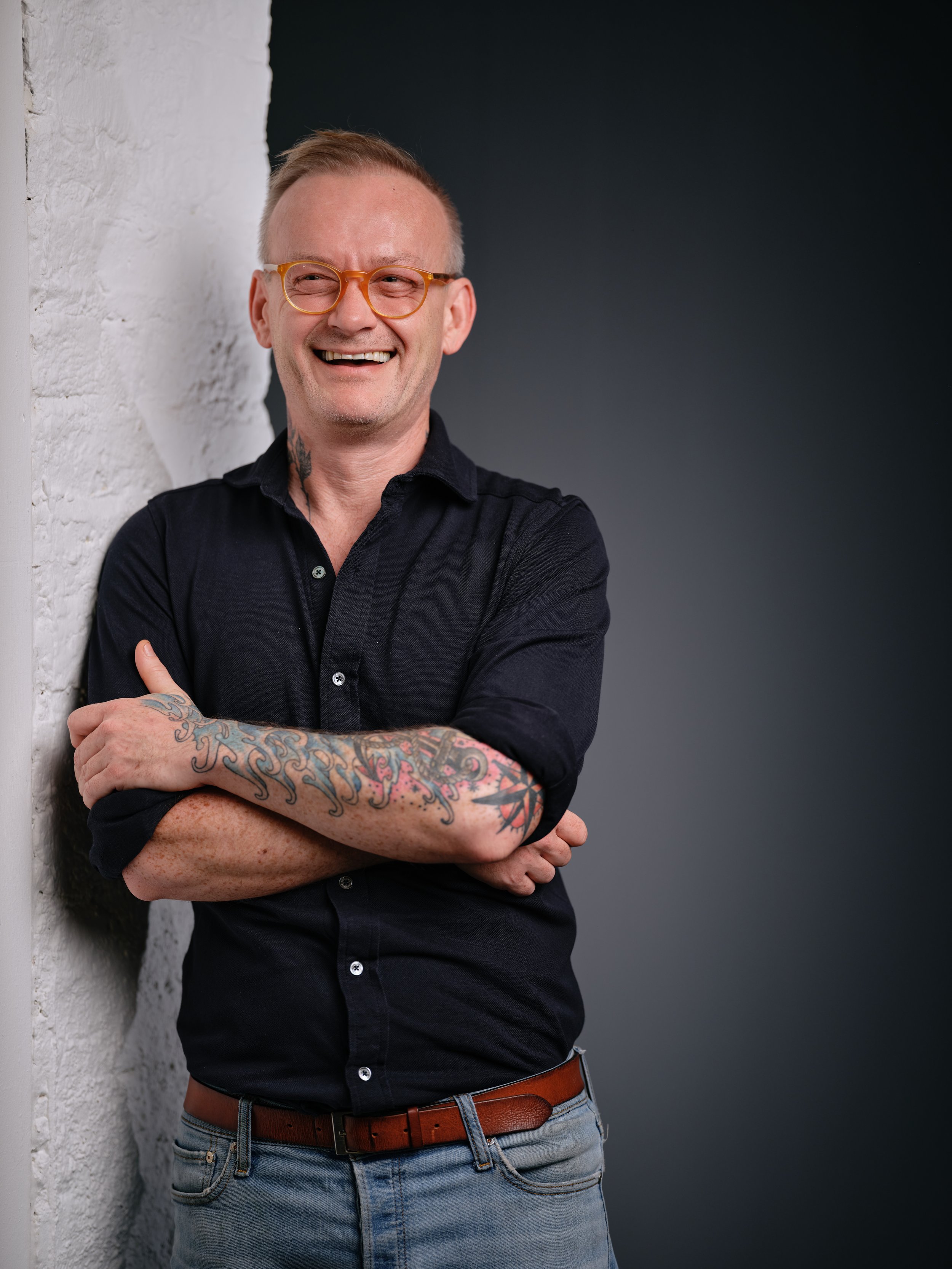 A man with glasses, short blonde hair, and tattoos on his arm, wearing a black shirt and jeans, smiling and leaning against a white textured wall with a dark grey background.