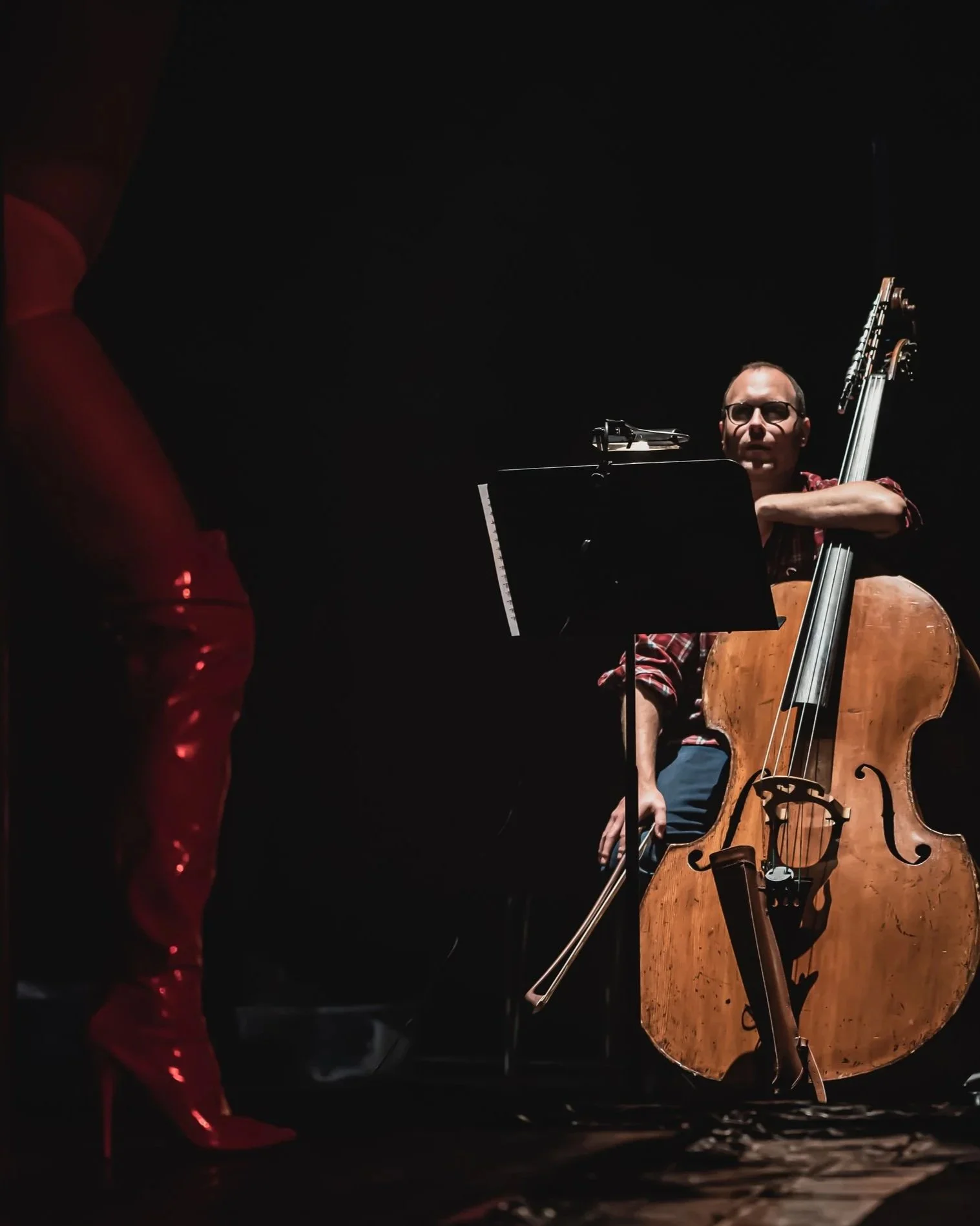 A man wearing glasses and a red plaid shirt playing a cello, with a music stand in front of him, on a dark stage. Part of another person in a red sparkling outfit is visible on the left.