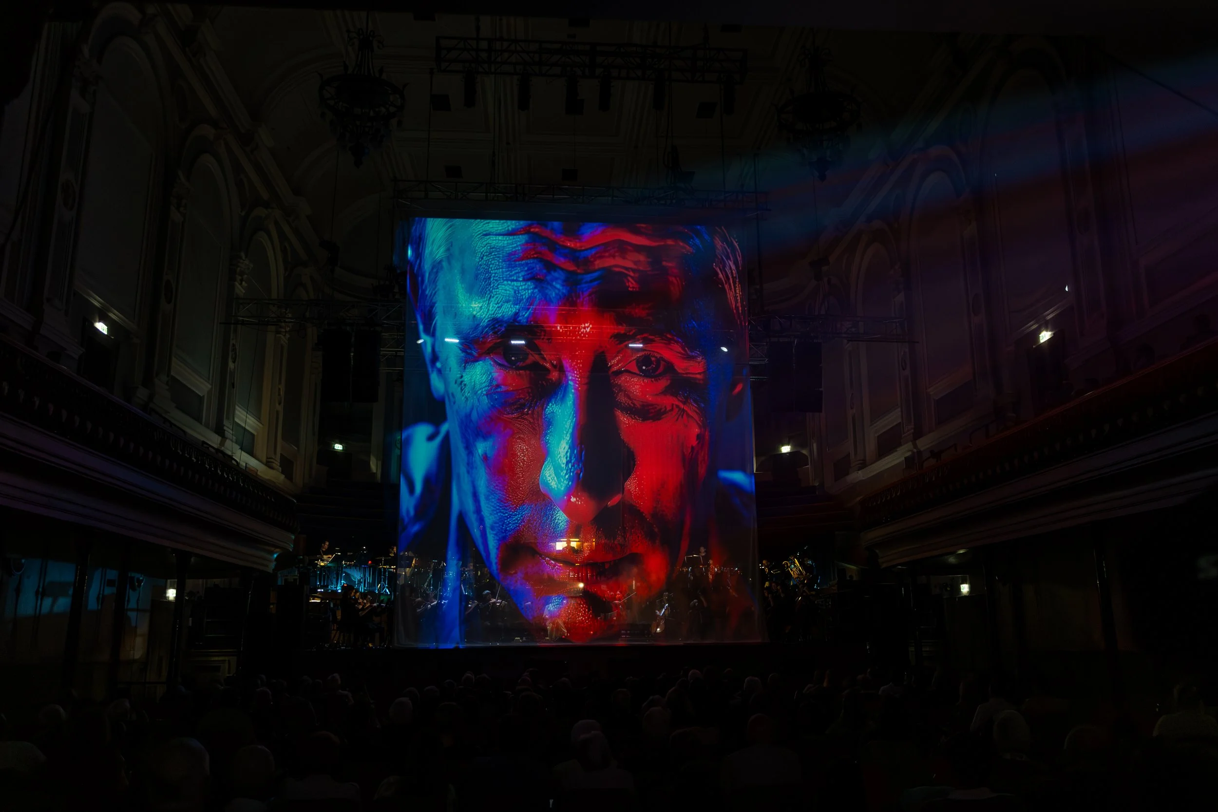 Large digital display shows a stylized face with vivid colors, including red, blue, and purple, in a grand hall with ornate architecture.