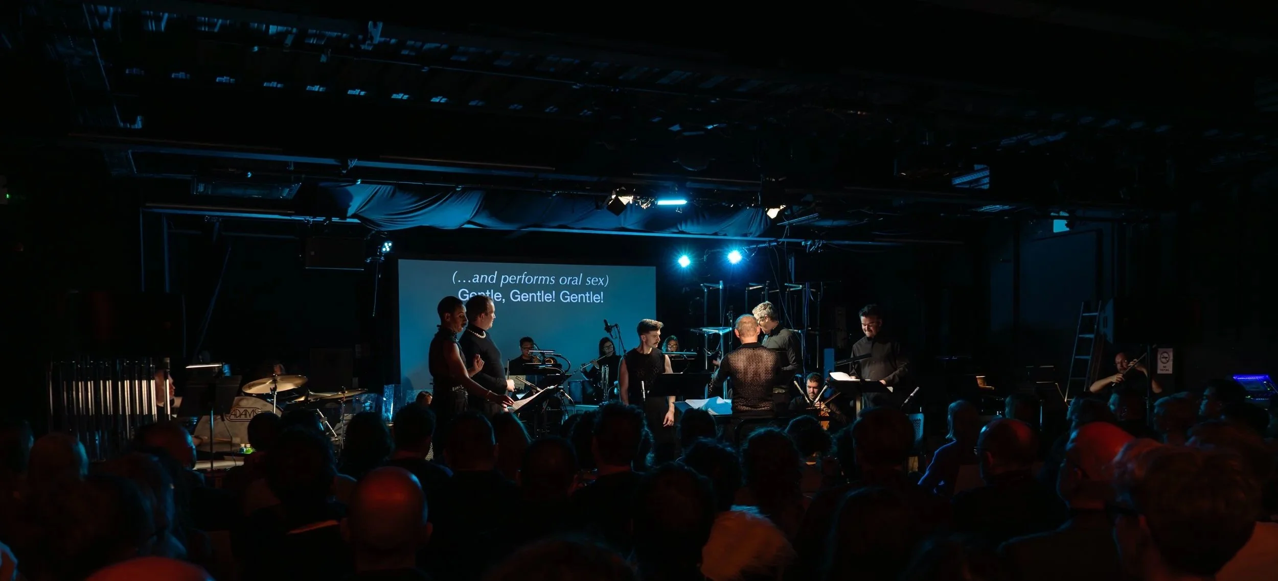 A stage performance with a group of people, some standing and some seated, in a dark concert venue. There are musicians, a large screen displaying lyrics, and an audience watching.