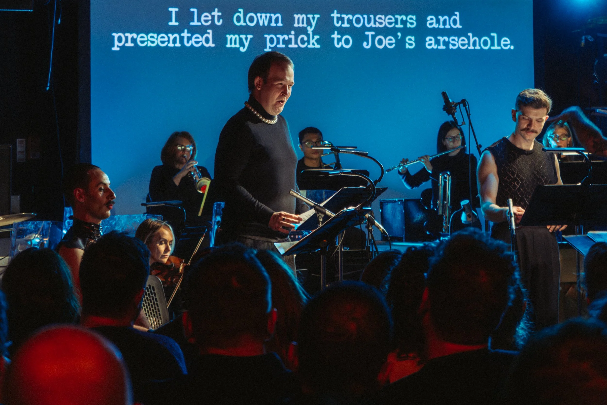 A group of musicians performing on stage with a large screen behind them displaying text, including a person in the foreground playing an instrument and others playing various instruments around him. Audience members are visible in the foreground.