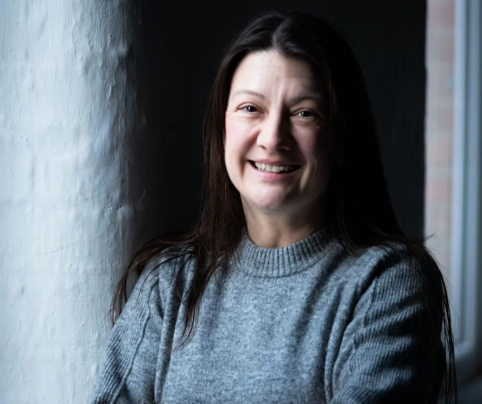 Smiling woman with long dark hair wearing a gray sweater, standing by a window with natural light, against a dark wall.