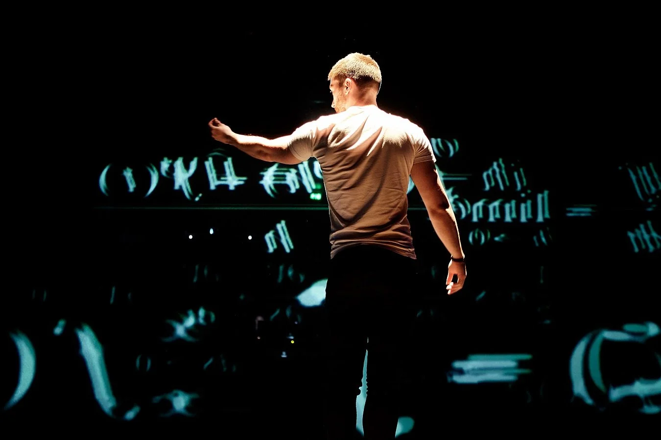 A person with short, blond hair and a beige t-shirt, standing on stage with a dark background featuring glowing blue and green digital symbols and numbers.