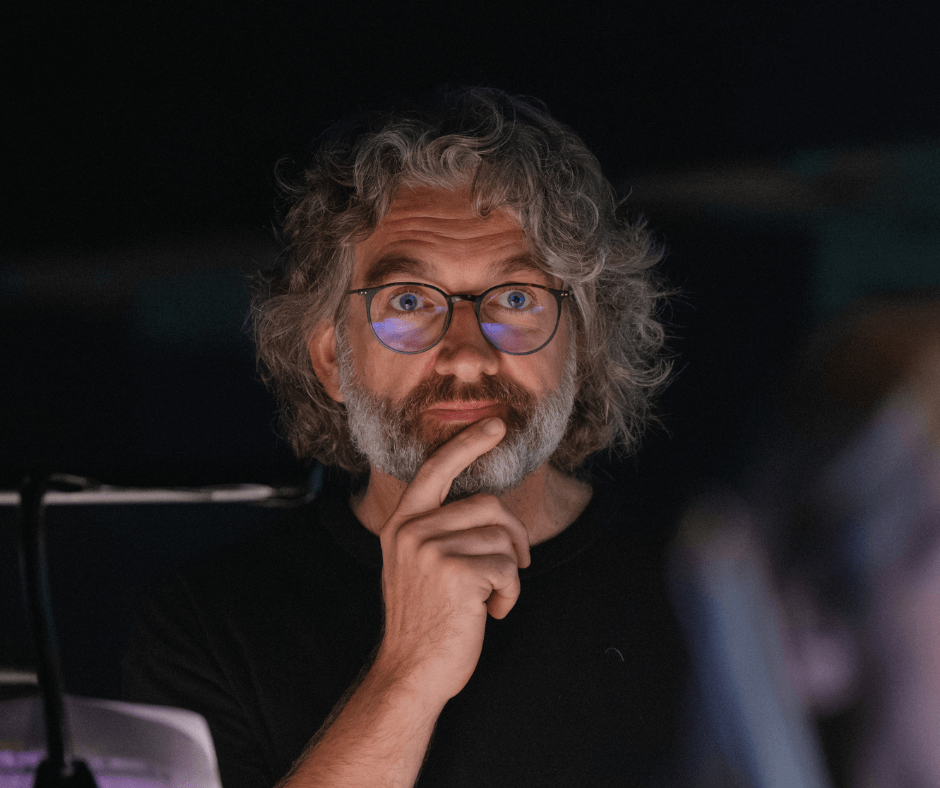 A man with curly gray hair and glasses, resting his chin on his hand, looking thoughtfully at something off-camera.