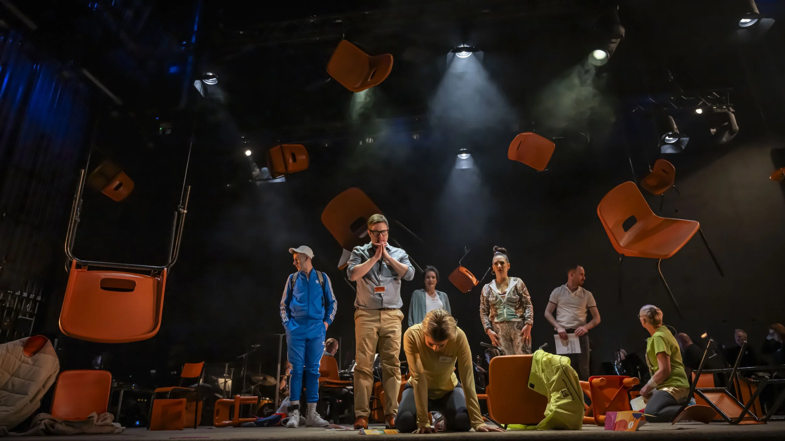 People on stage preparing for a performance, with orange chairs suspended from the ceiling and stage lights overhead.