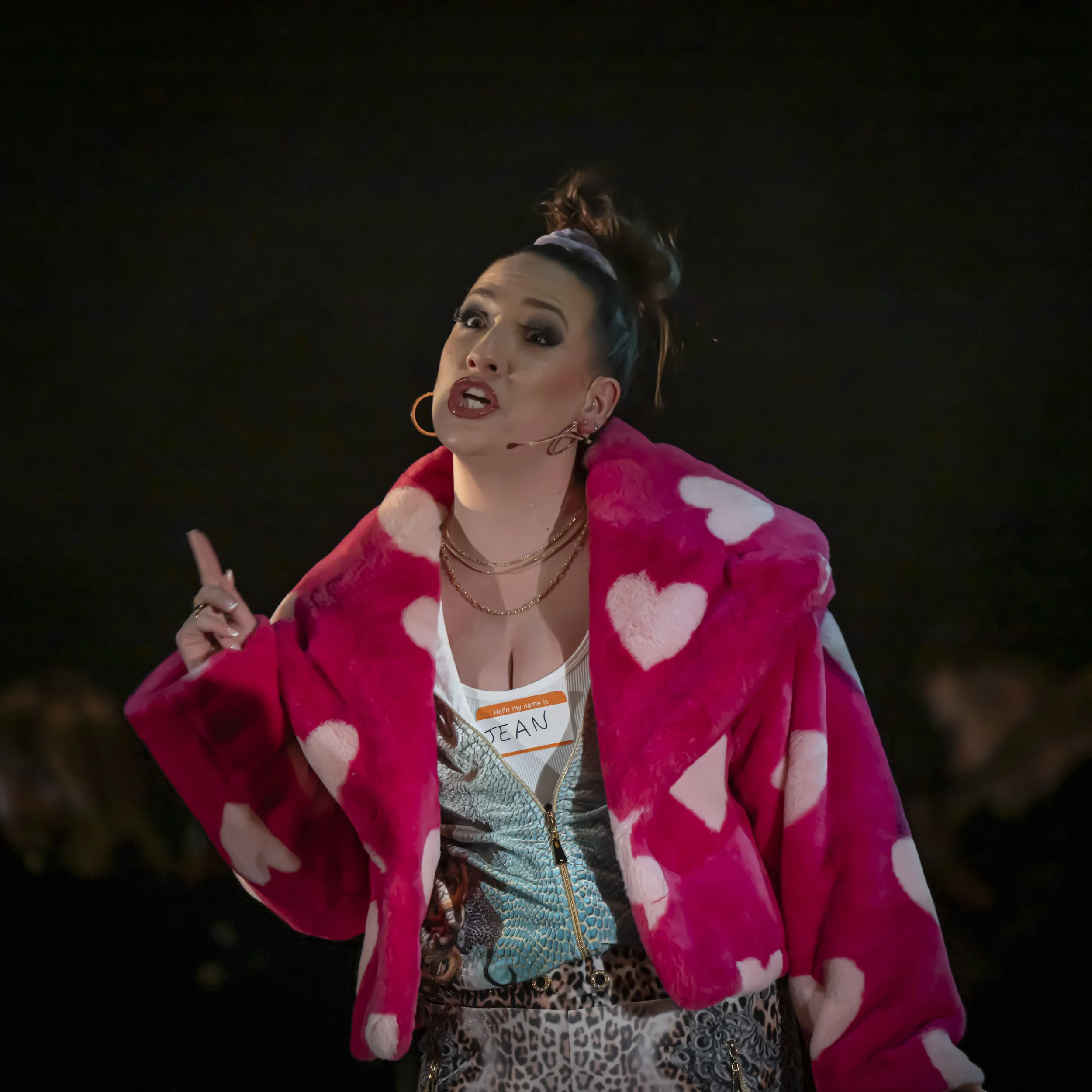 A woman with styled hair, makeup, and jewelry is wearing a pink and white fuzzy jacket over a patterned dress, and has a name tag that reads "JEAN," while speaking and gesturing with her hand against a dark background.